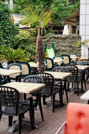 Terrasse mit Tischen und Stühlen, umgeben von Grünpflanzen und einer Steinmauer.