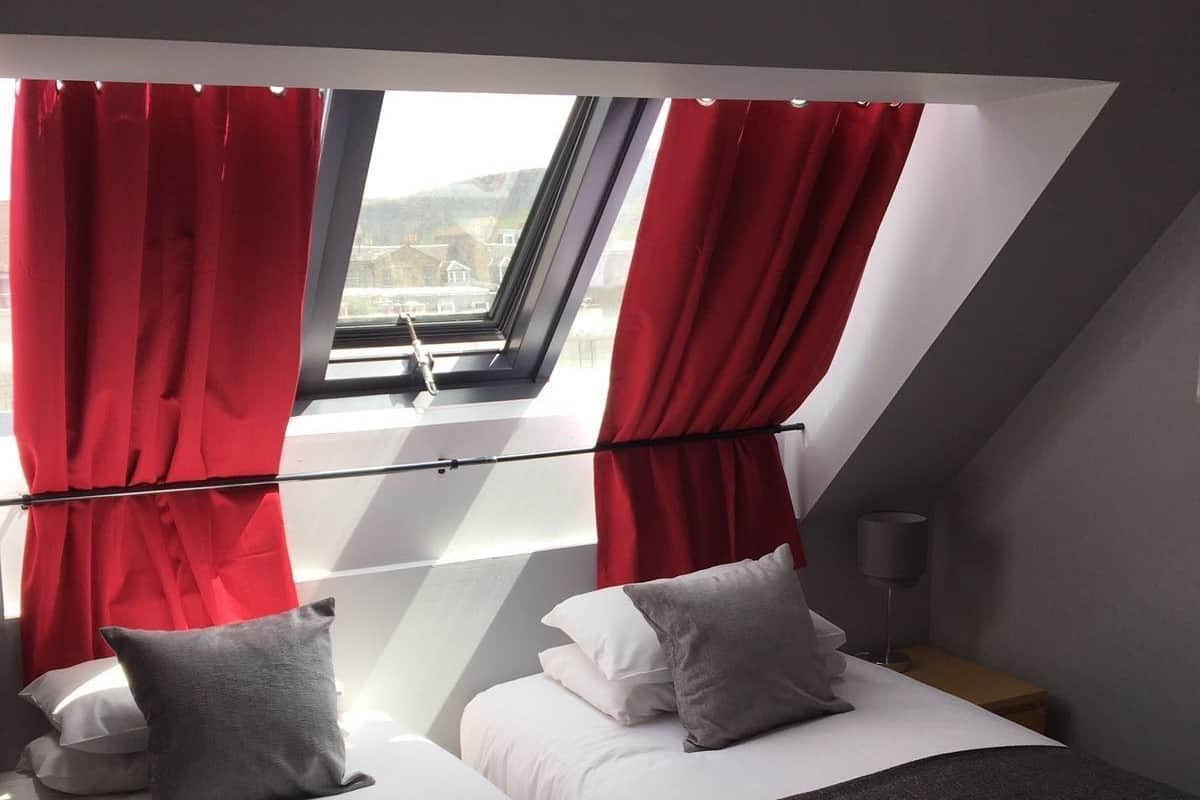 Une chambre avec deux lits, des rideaux rouges, une lucarne et une vue sur la ville.