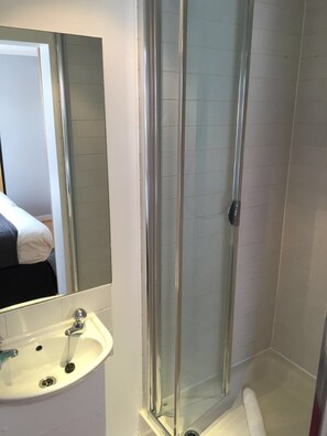 A bathroom with a glass-enclosed shower, a white sink with a mirror, and a bed visible in the background.