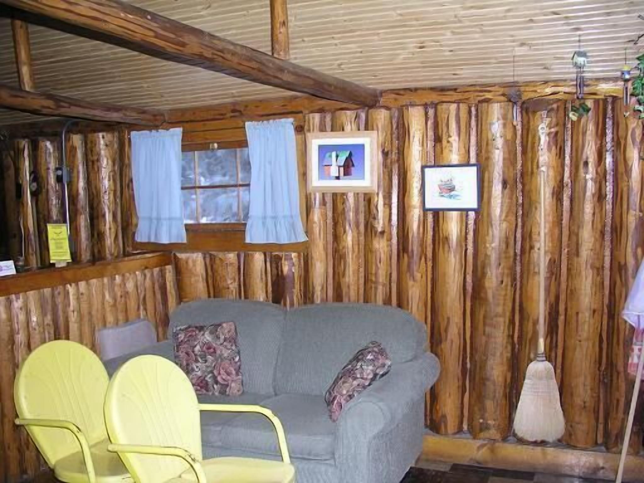 A rustic cabin interior with a wooden wall, a gray sofa, two yellow chairs, and a broom.