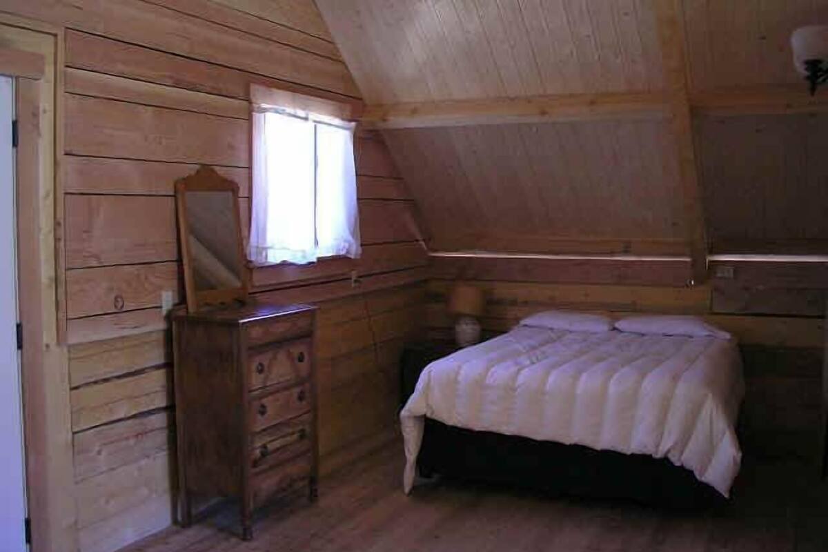 Un dormitorio de cabaña de madera con cama, cómoda, espejo y ventana.
