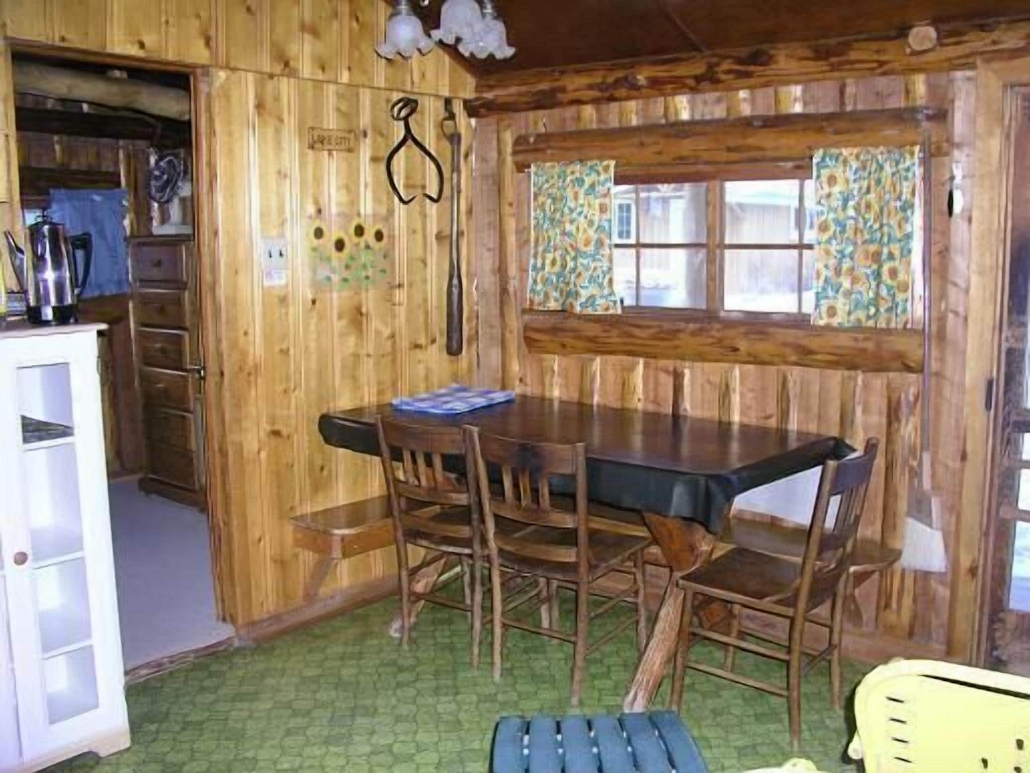 Interior de madera con una mesa y sillas de comedor, un banco, una ventana con cortinas y una zona de cocina con armarios y un lavamanos.