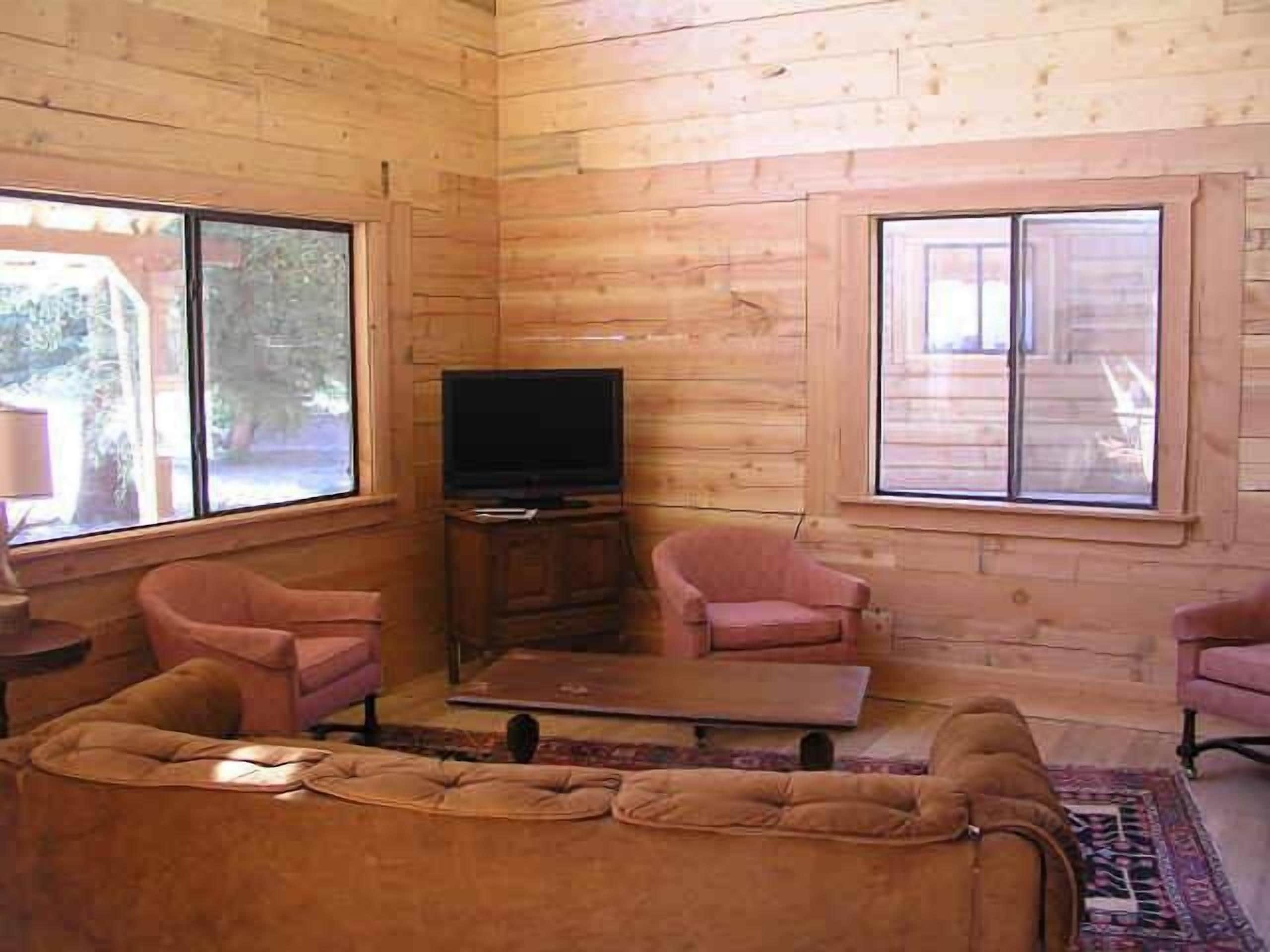 L’intérieur d’un chalet en bois comprend un canapé, deux fauteuils, un téléviseur posé sur un meuble en bois et de grandes fenêtres.