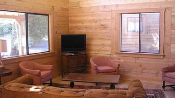 L’intérieur d’un chalet en bois comprend un canapé, deux fauteuils, un téléviseur posé sur un meuble en bois et de grandes fenêtres.