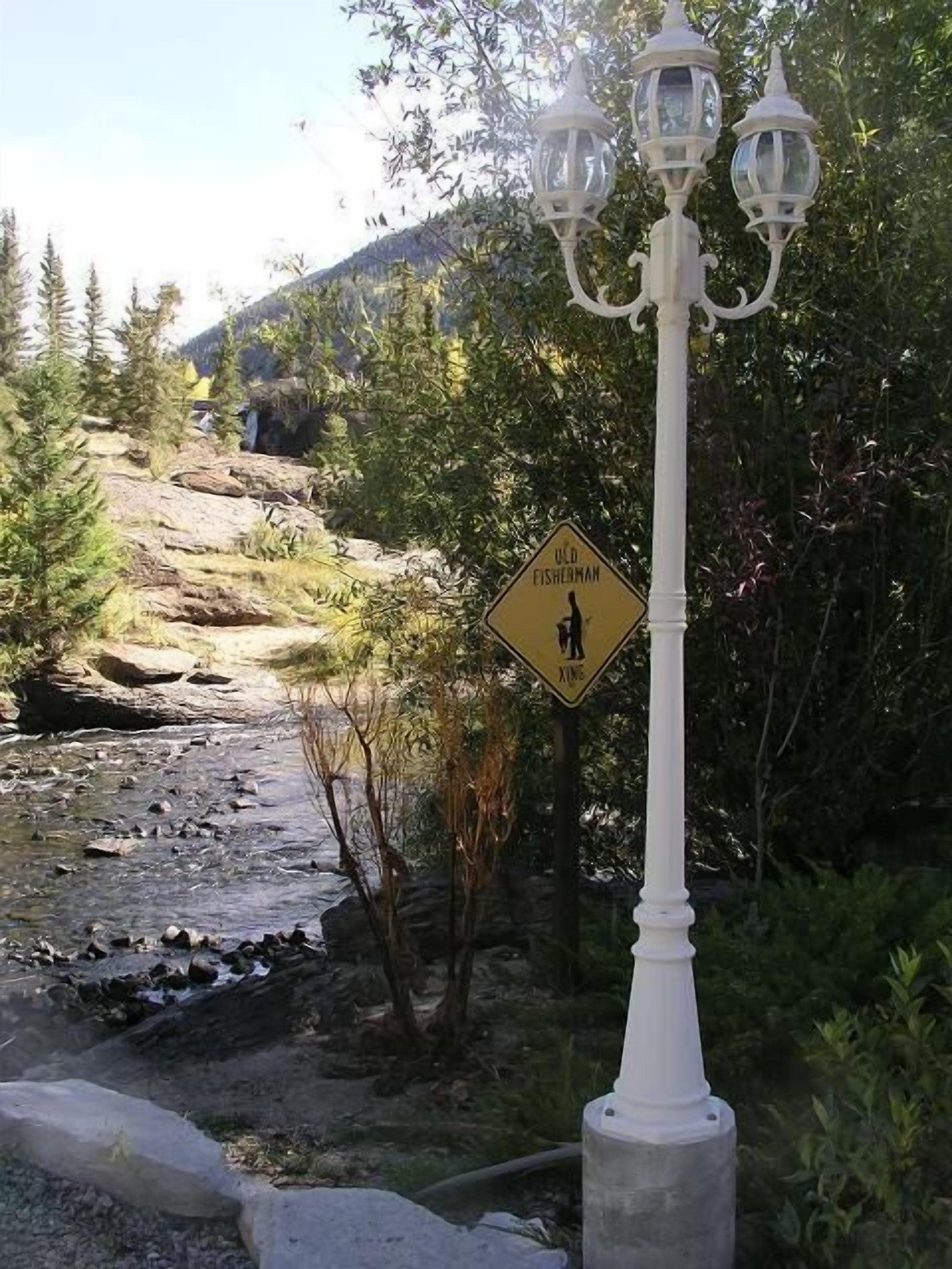 Señal de paso para peatones cerca de un arroyo pedregoso con un farol blanco y una montaña al fondo.
