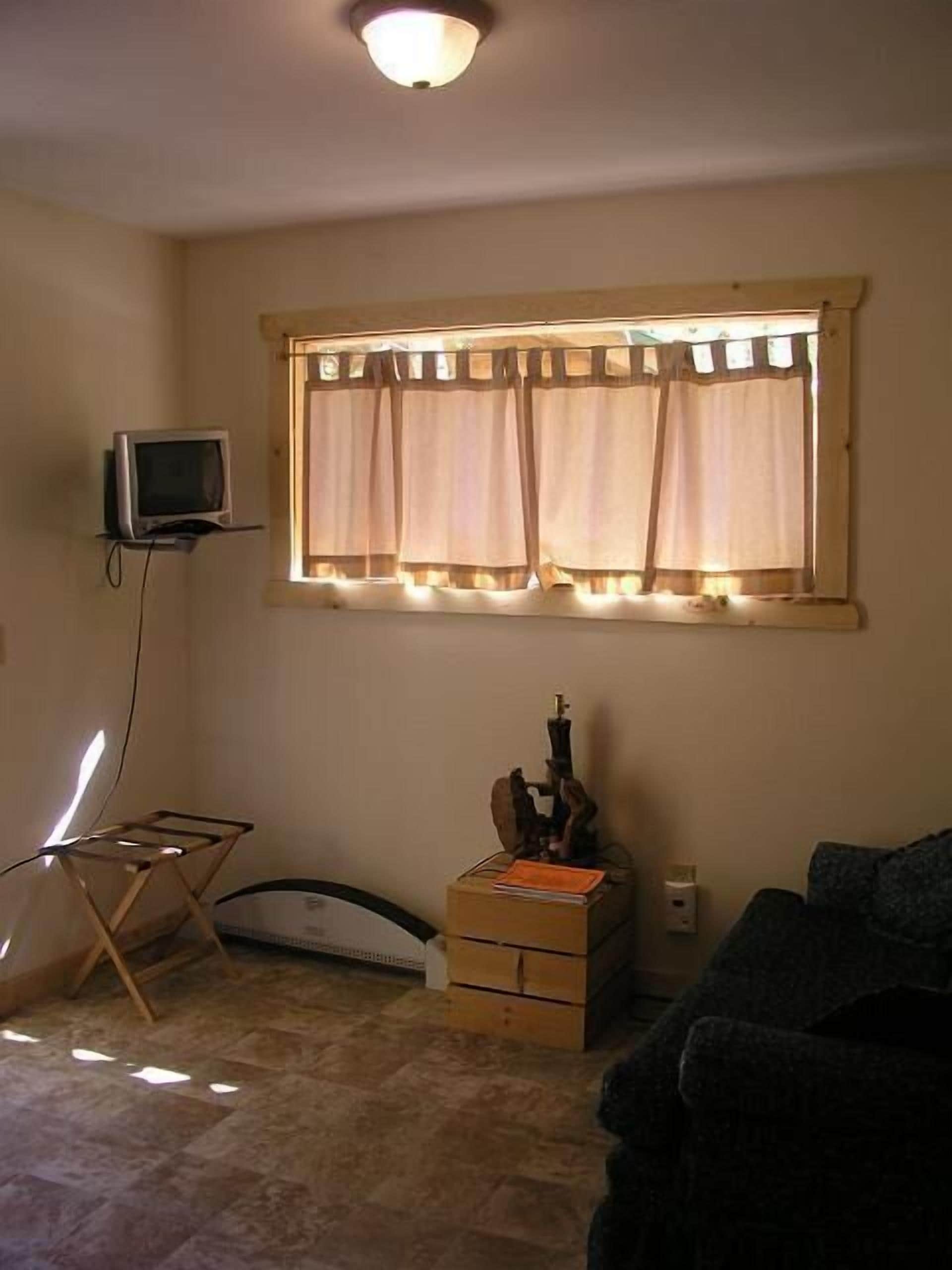 Una habitación con televisión sobre un mueble bajo, una ventana con cortinas, una mesita con una guitarra y un sofá.