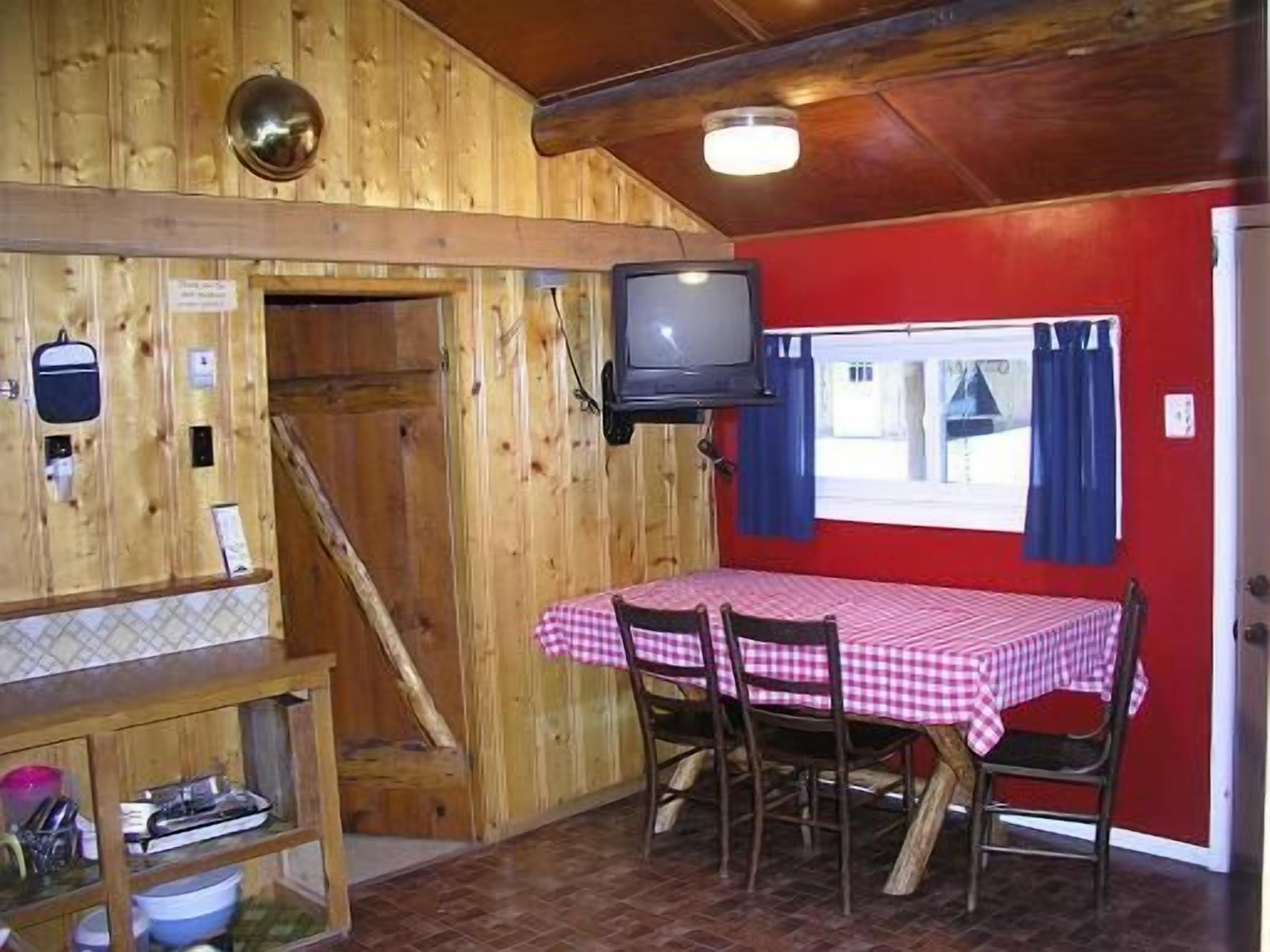Un interior de madera con una mesa de comedor preparada para cuatro personas, un televisor montado en la pared y un ventanal con cortinas.