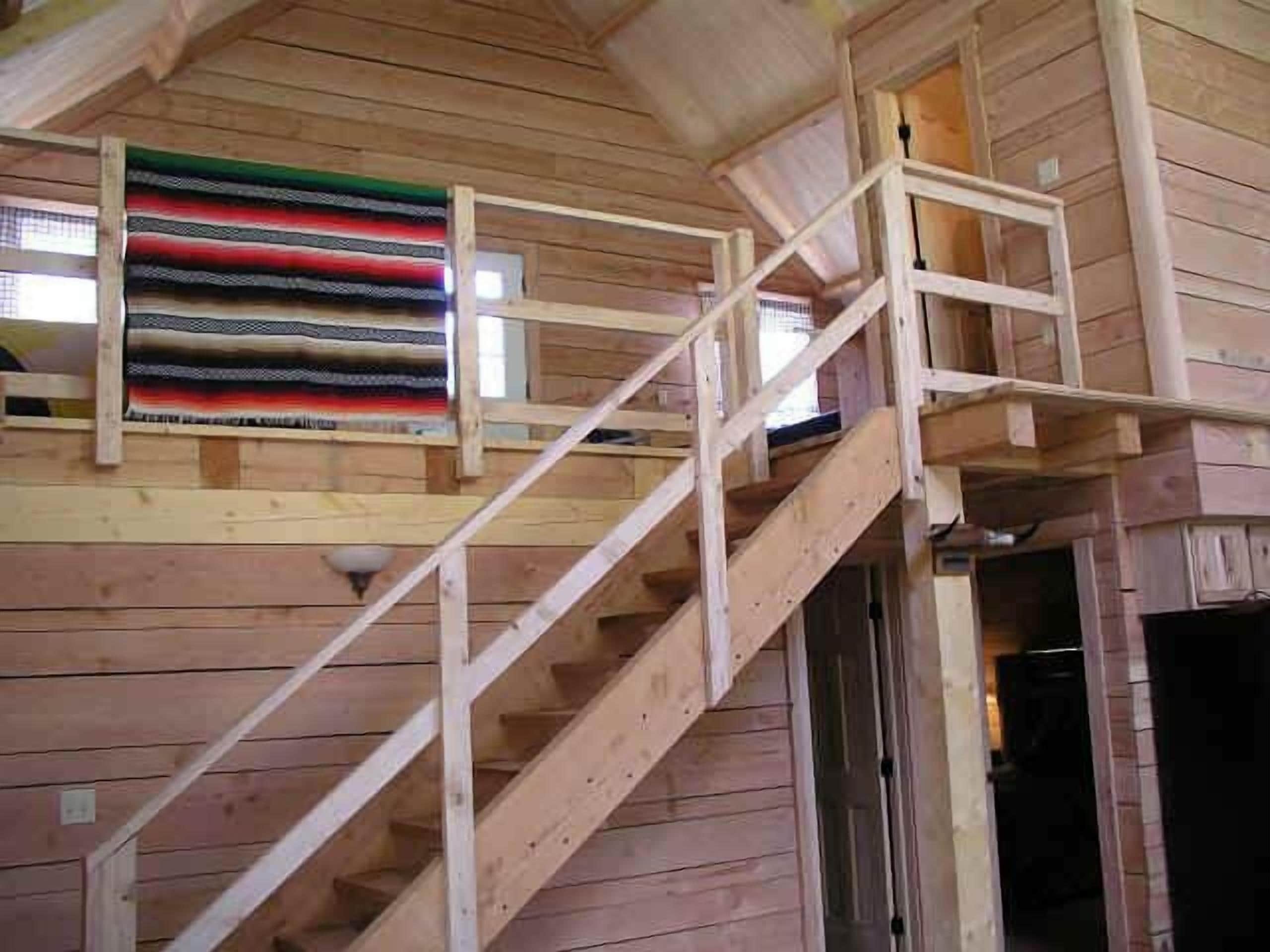 Una escalera de madera que sube a un altillo con un ventanal que da a un jardín.