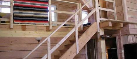 Una escalera de madera que sube a un altillo con un ventanal que da a un jardín.