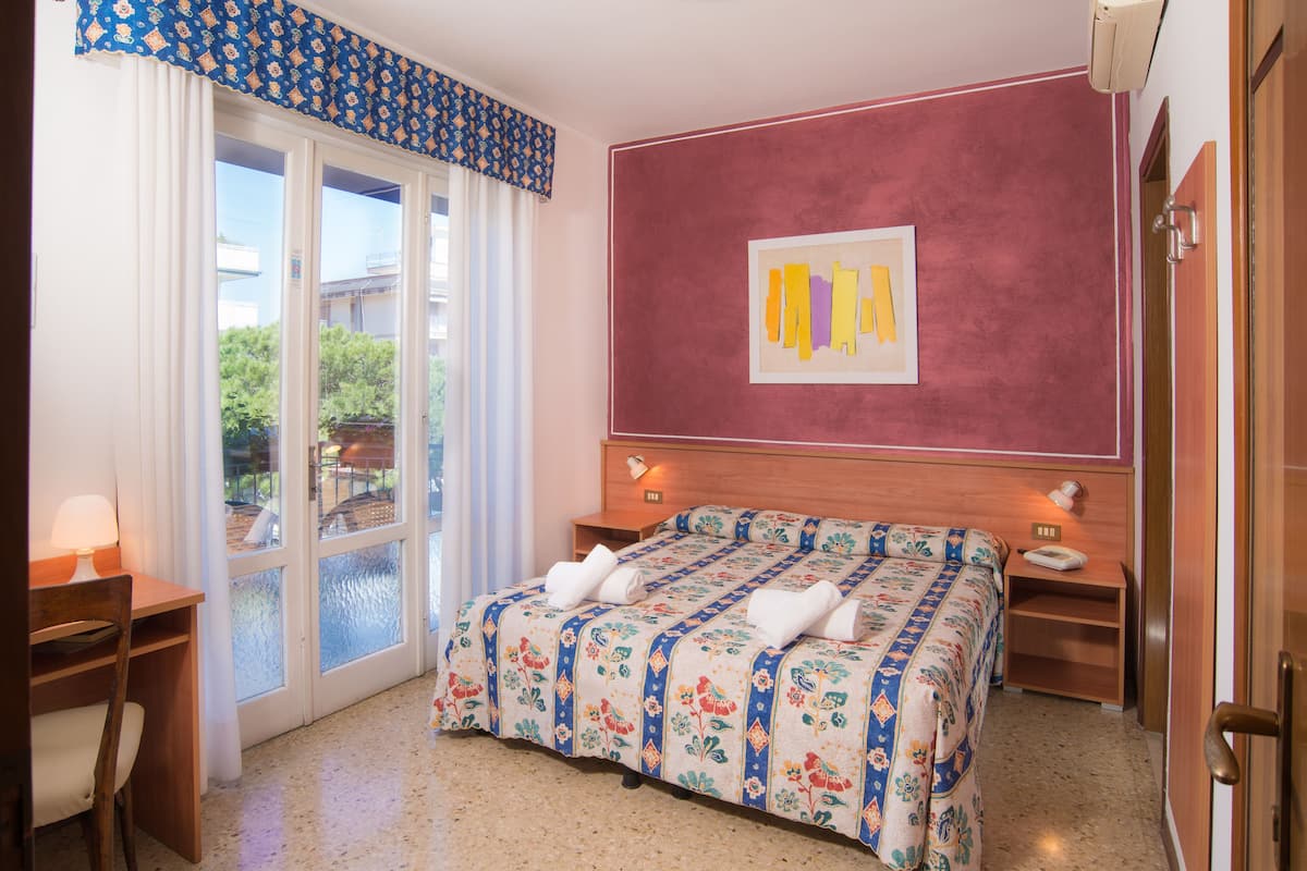 A bedroom with a double bed, a desk, a chair, a nightstand, and a window with a view of greenery.