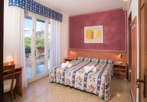 A bedroom with a double bed, a desk, a chair, a nightstand, and a window with a view of greenery. - Hotel Azzorre & Antille (Jesolo)