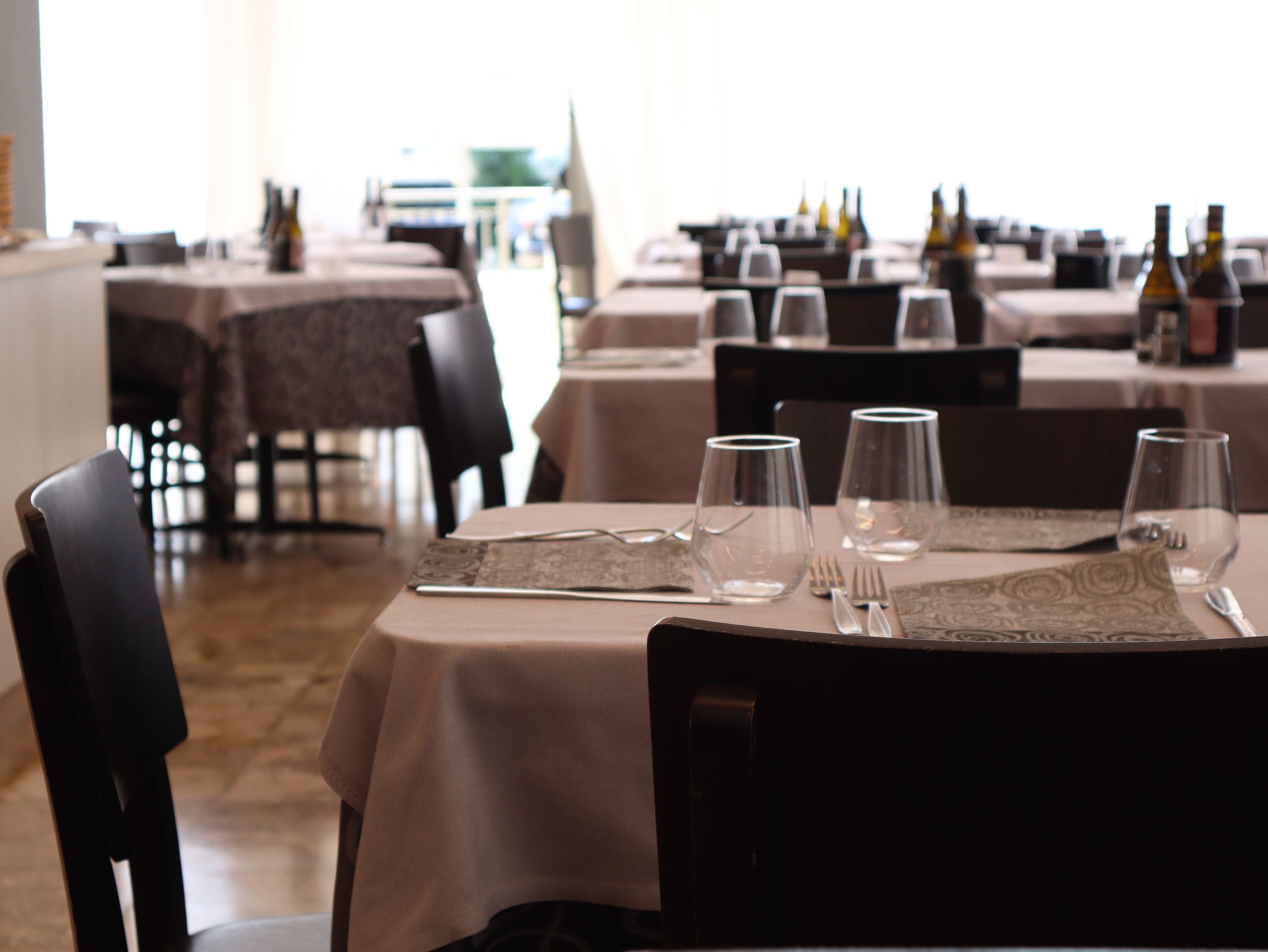 Een restaurant met gedekte tafels, stoelen en wijnflessen op de achtergrond.