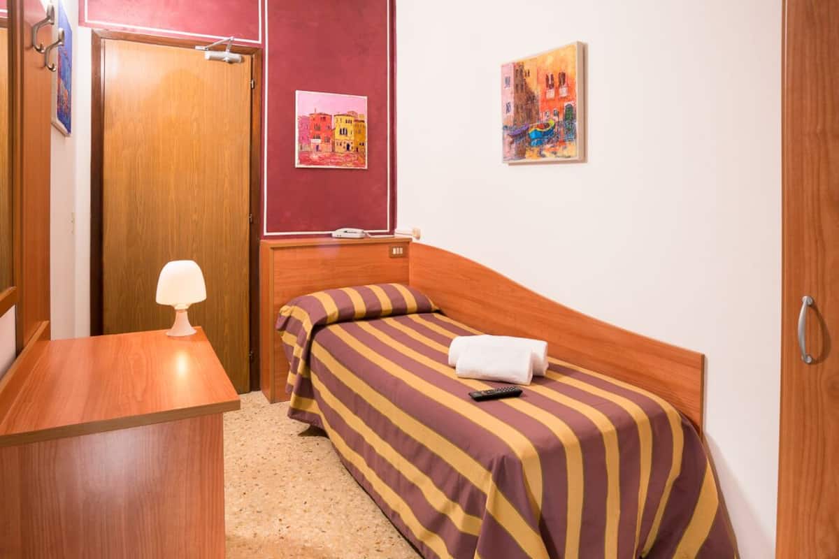 A small, neatly arranged room with a wooden bed, a bedside table with a lamp, and two paintings on the wall.