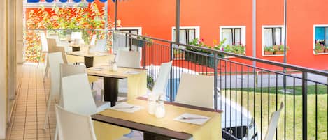 An outdoor dining area with tables and chairs, a railing, and a view of a red building with windows and plants.