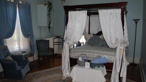 A four-poster canopy bed with a white bedspread, a chair, a table with a basket, and a lamp.
