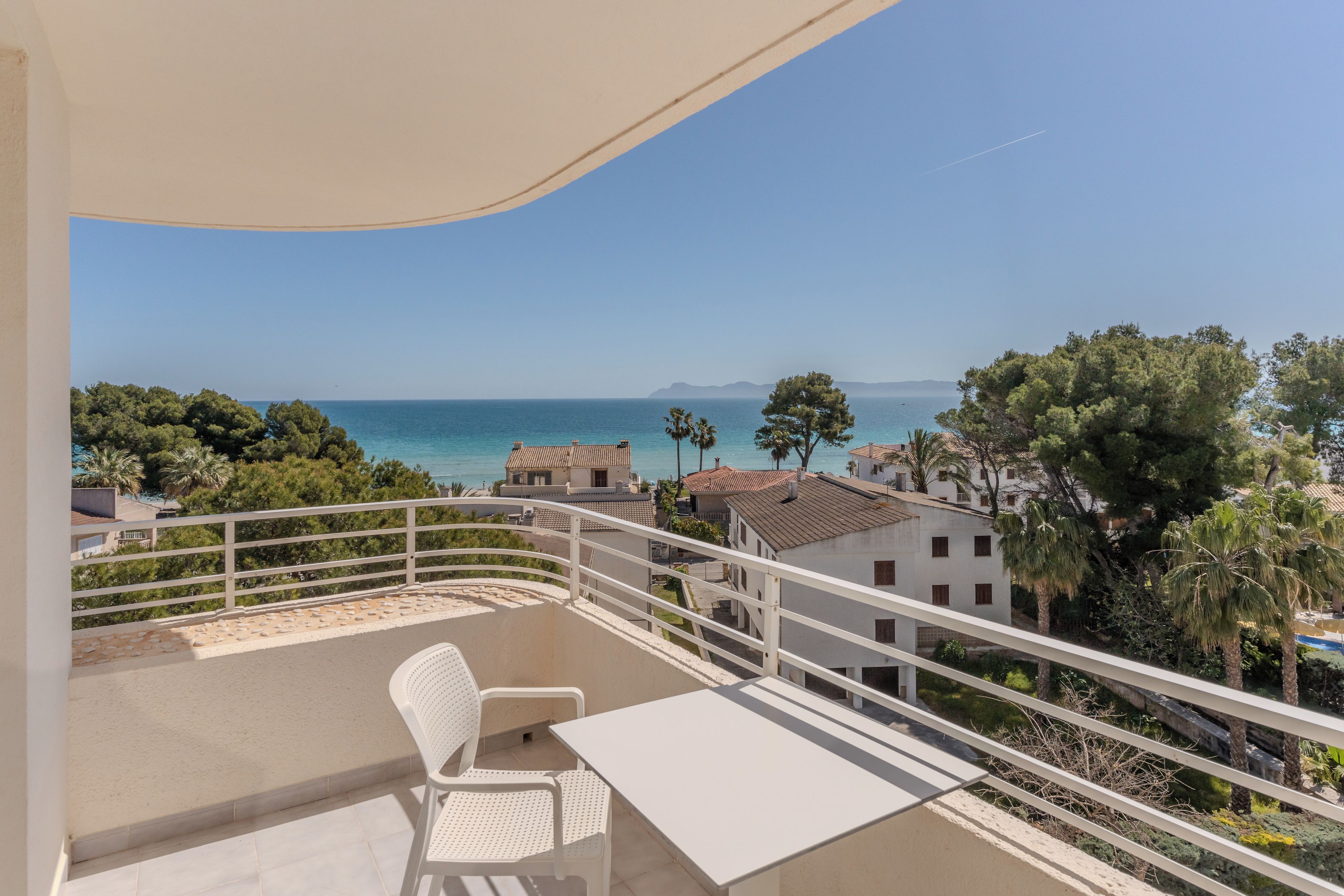 A balcony with a white table and chair, overlooking a coastal view with buildings and trees.