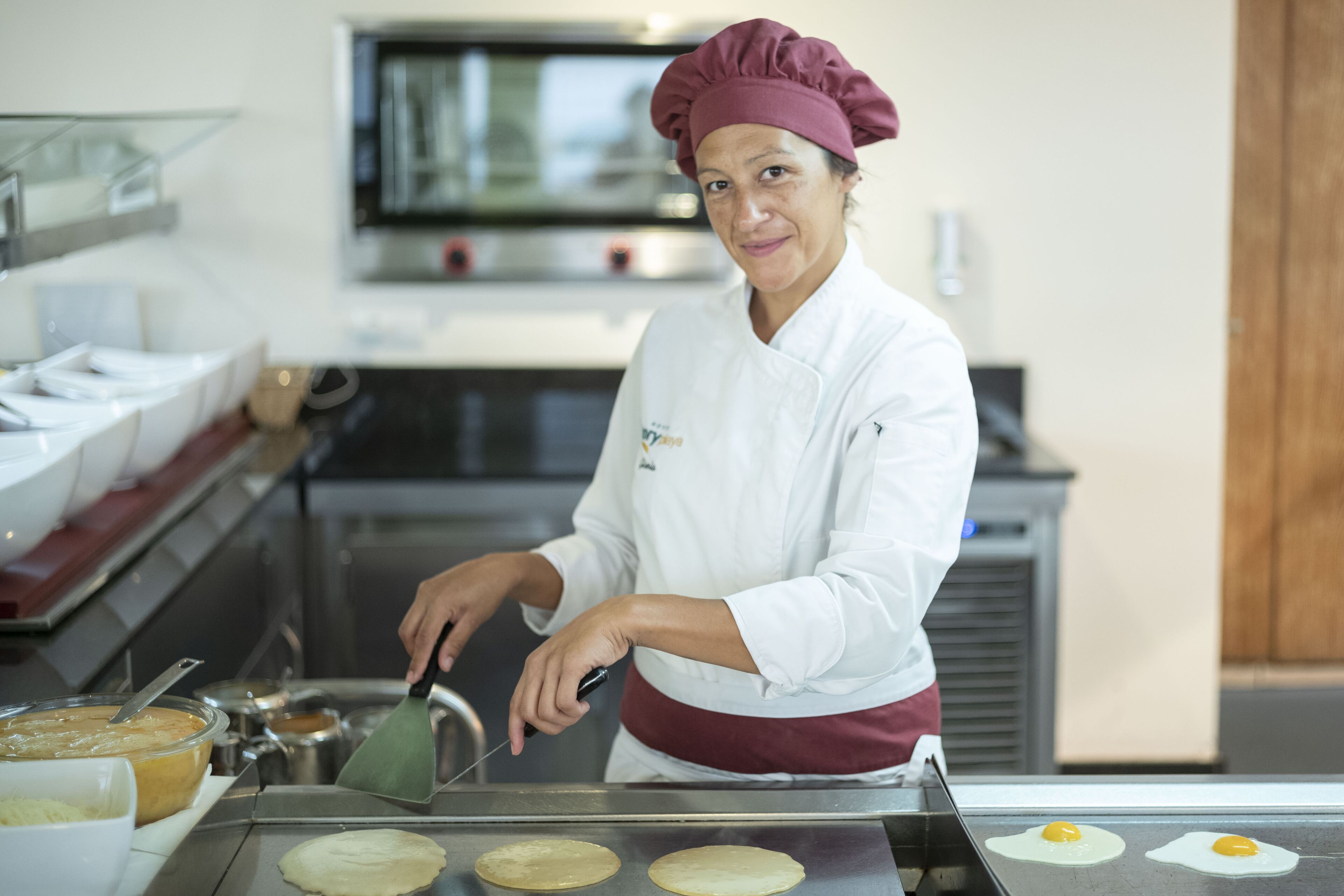 A chef in a white uniform and maroon hat cooking on a griddle in a kitchen.