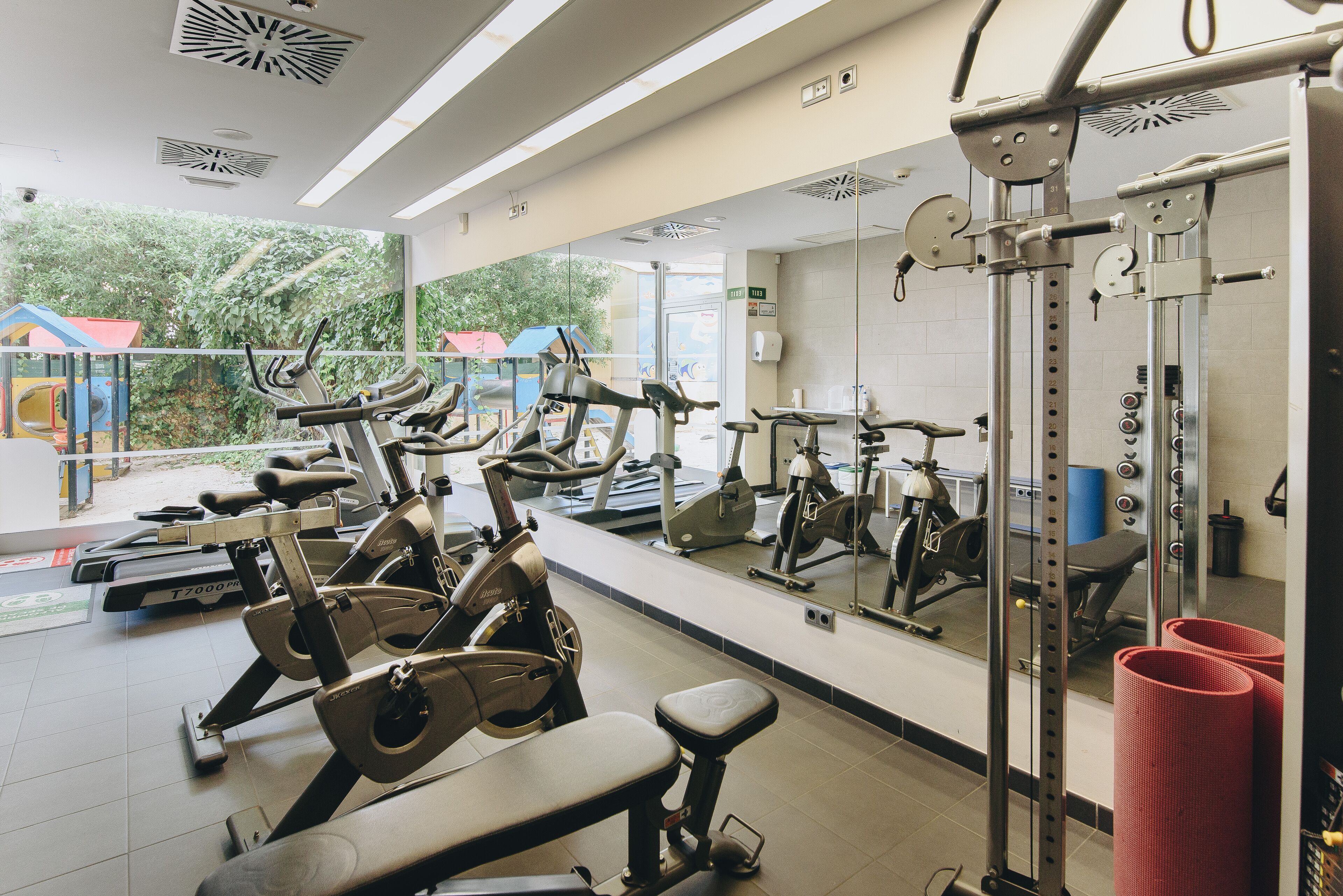 Une salle de sport équipée de divers appareils d’exercice, dotée d’un grand miroir et offrant une vue sur l’aire de jeux extérieure.