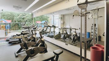 Une salle de sport équipée de divers appareils d’exercice, dotée d’un grand miroir et offrant une vue sur l’aire de jeux extérieure.