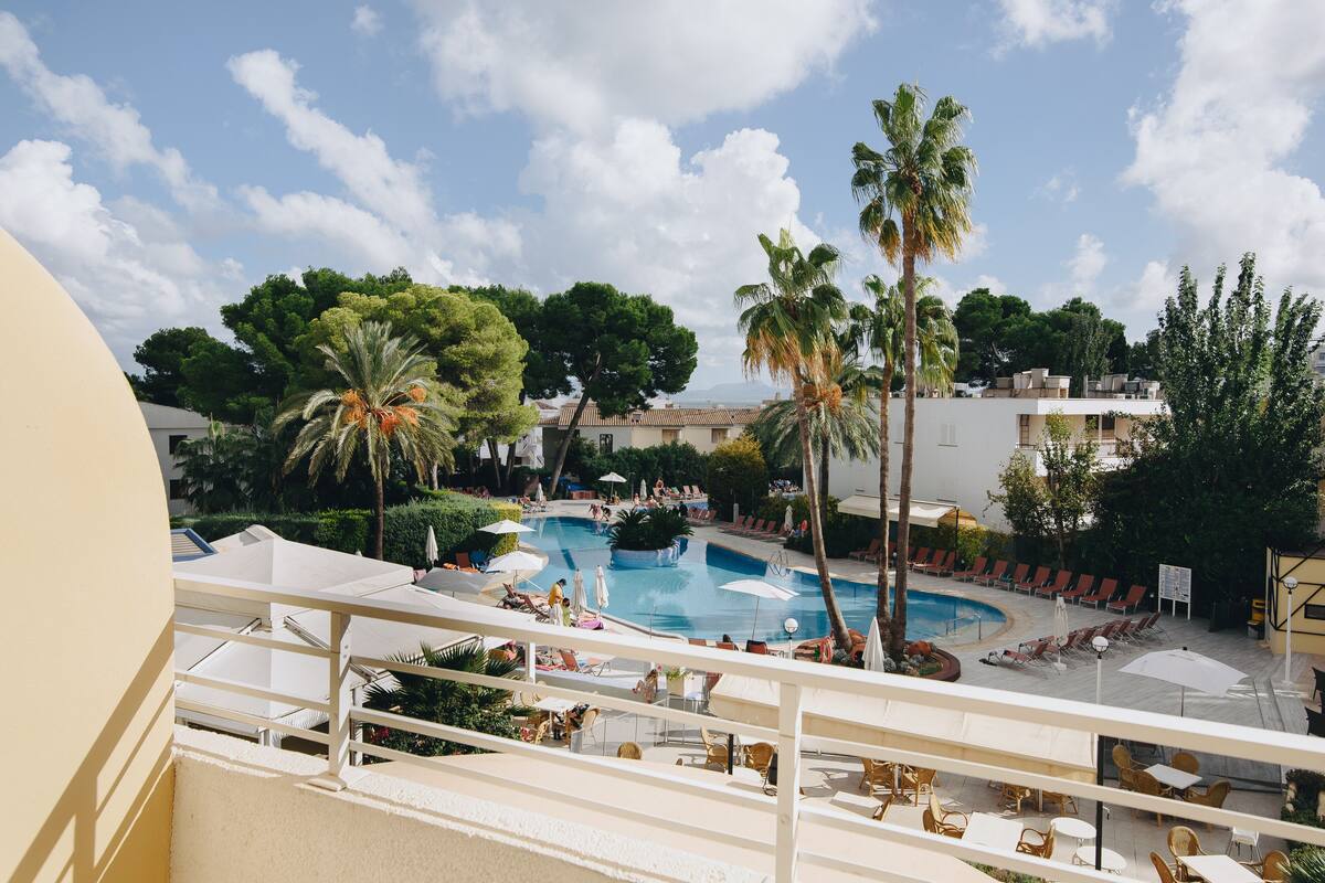 Un espace piscine doté de chaises longues et de parasols, entouré de palmiers et d’immeubles.