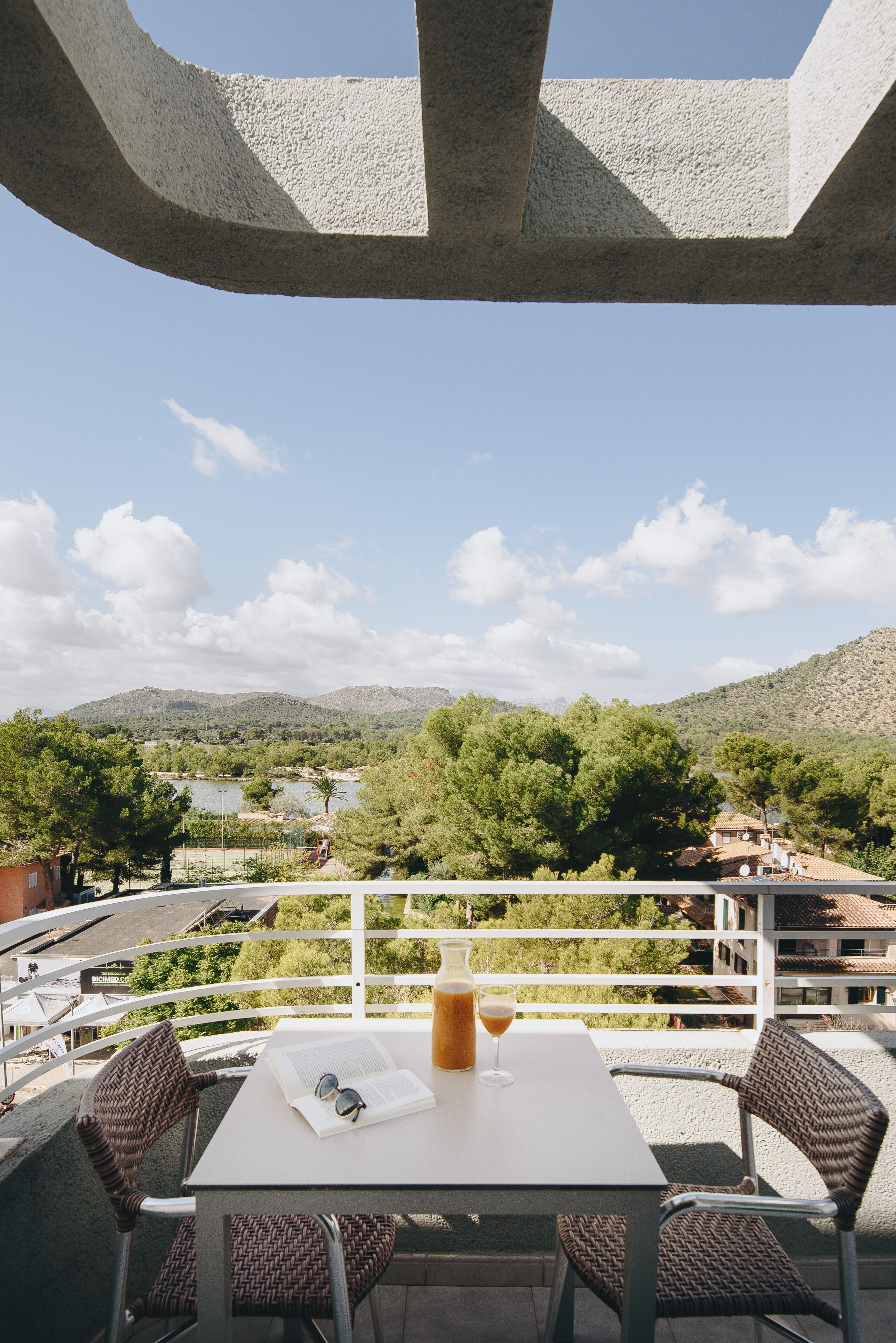 A terrace with a table, chairs, and a drink, overlooking a mountainous landscape with trees and buildings.
