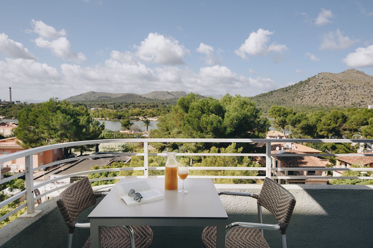 Un balcon avec une table, des chaises et une boisson. Le balcon offre une vue panoramique sur les montagnes et une ville.