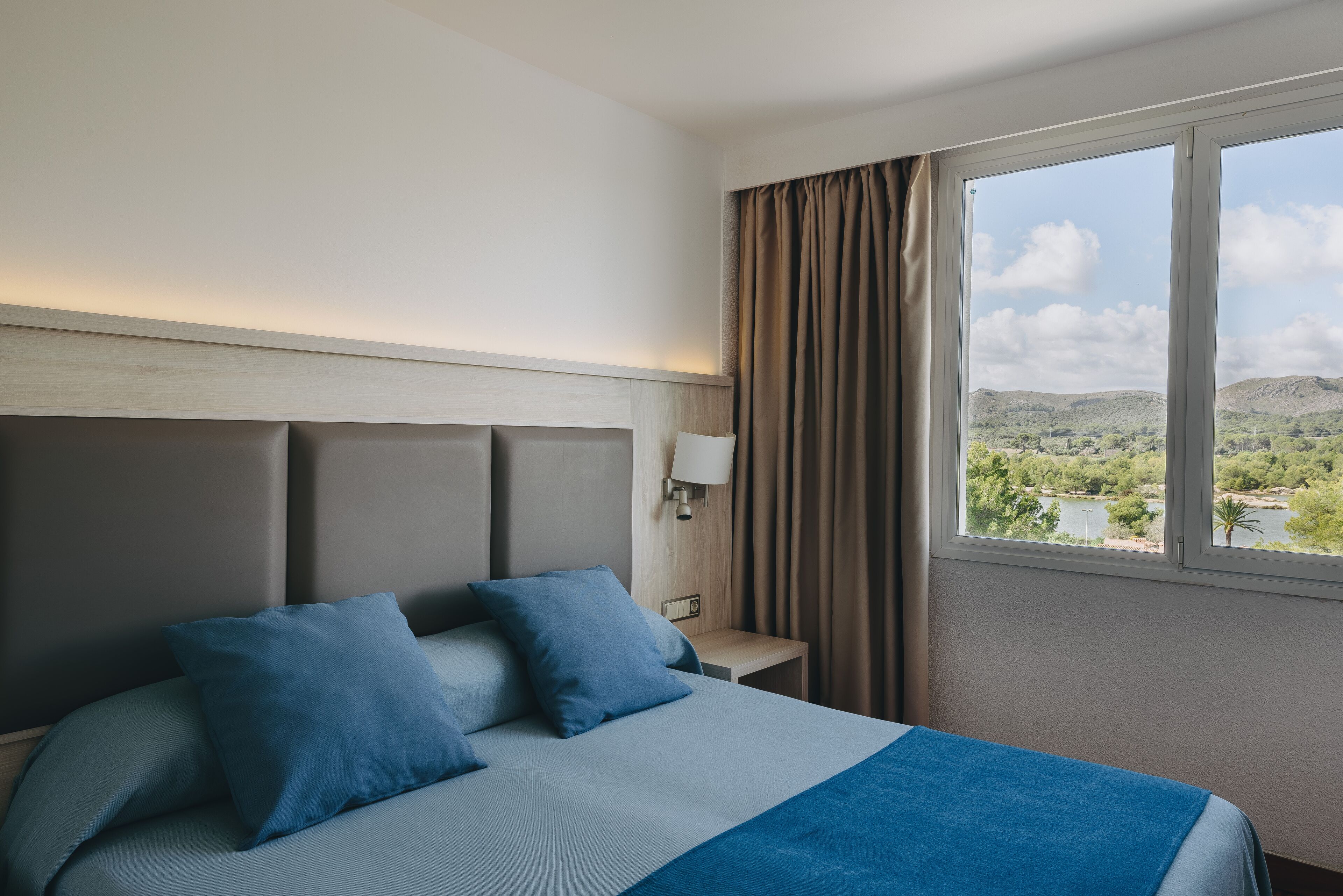 A hotel room with a bed, a nightstand, a lamp, and a window with a view of a landscape.