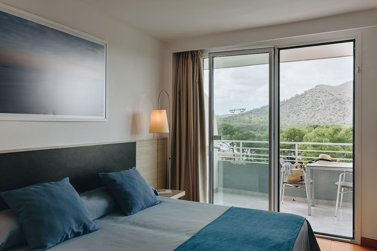 Une chambre d’hôtel avec un lit, une table de chevet, une lampe, une vue sur un balcon avec une table et des chaises, et un tableau accroché au mur.