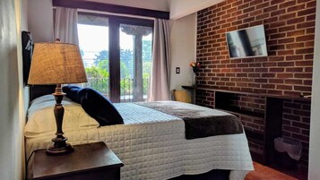 Un dormitorio con cama, lámpara, televisor montado en una pared de ladrillo y ventana con cortinas.