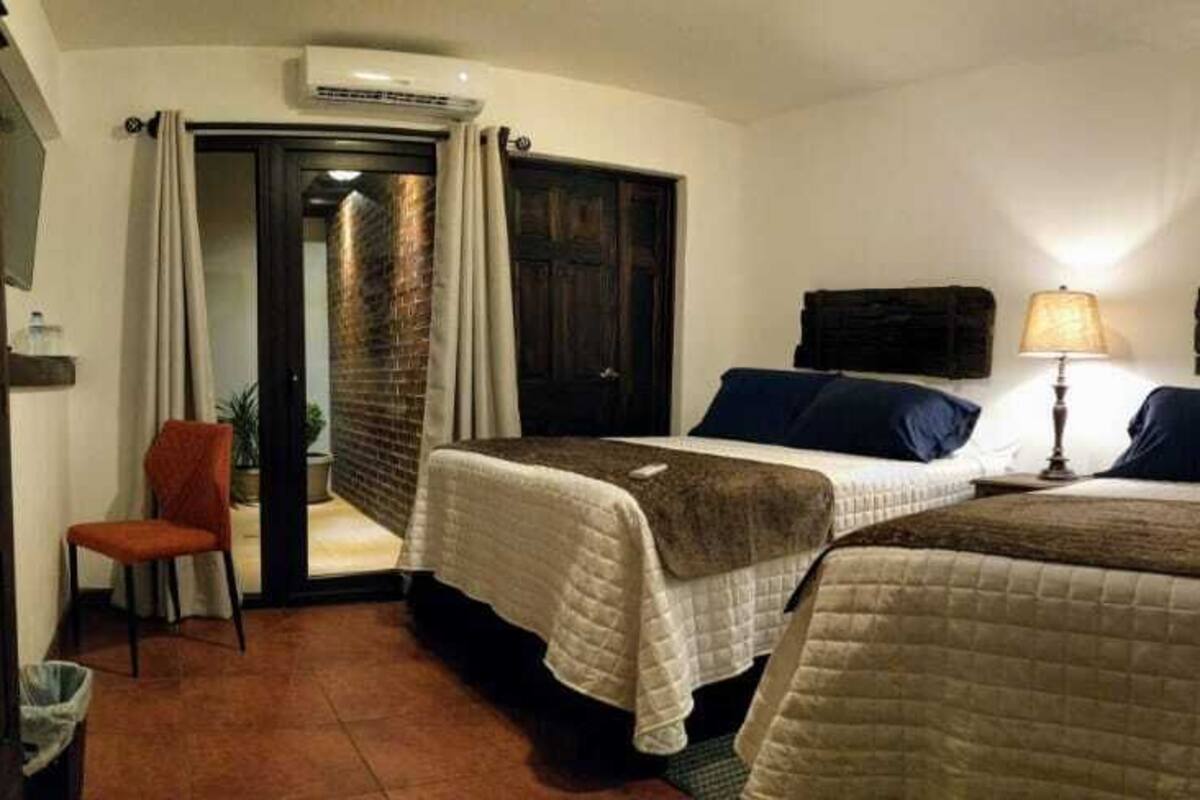A hotel room with two beds, a chair, a lamp, and a door leading to a corridor.