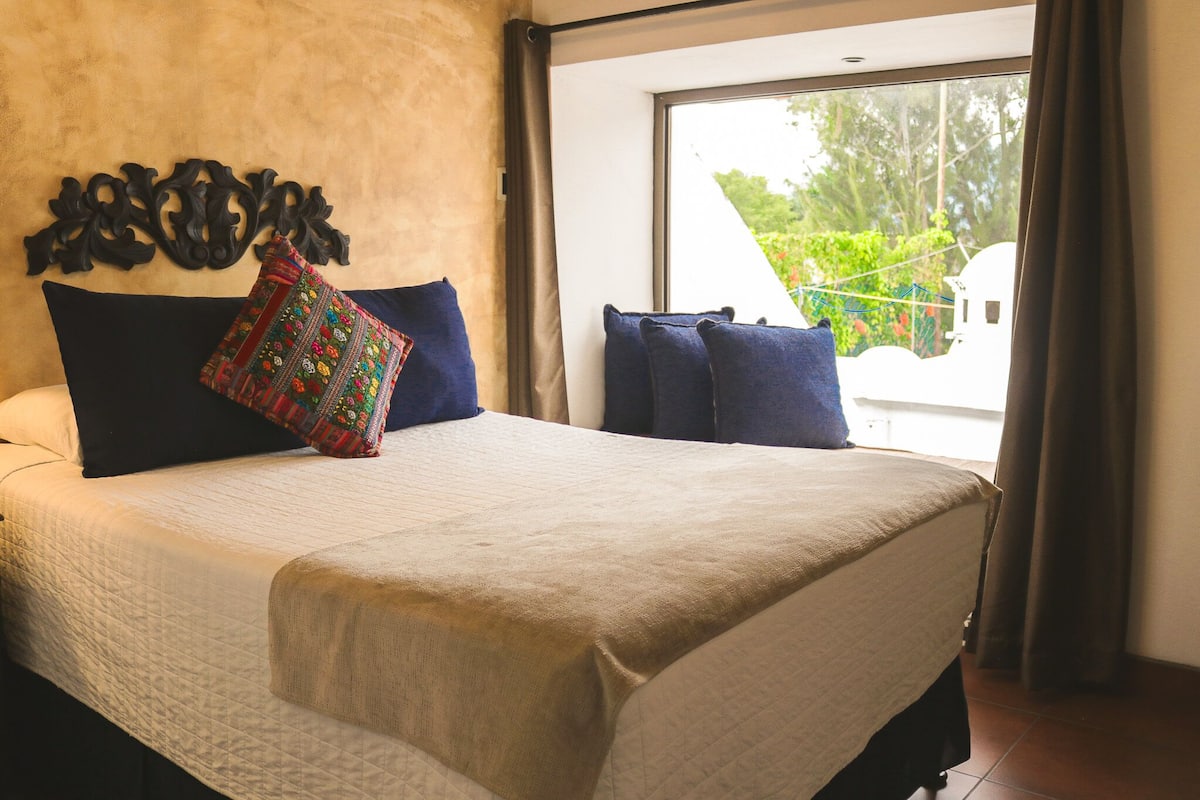 A neatly made bed with a beige bedspread and blue pillows, a decorative wall piece above it, and a window with curtains.