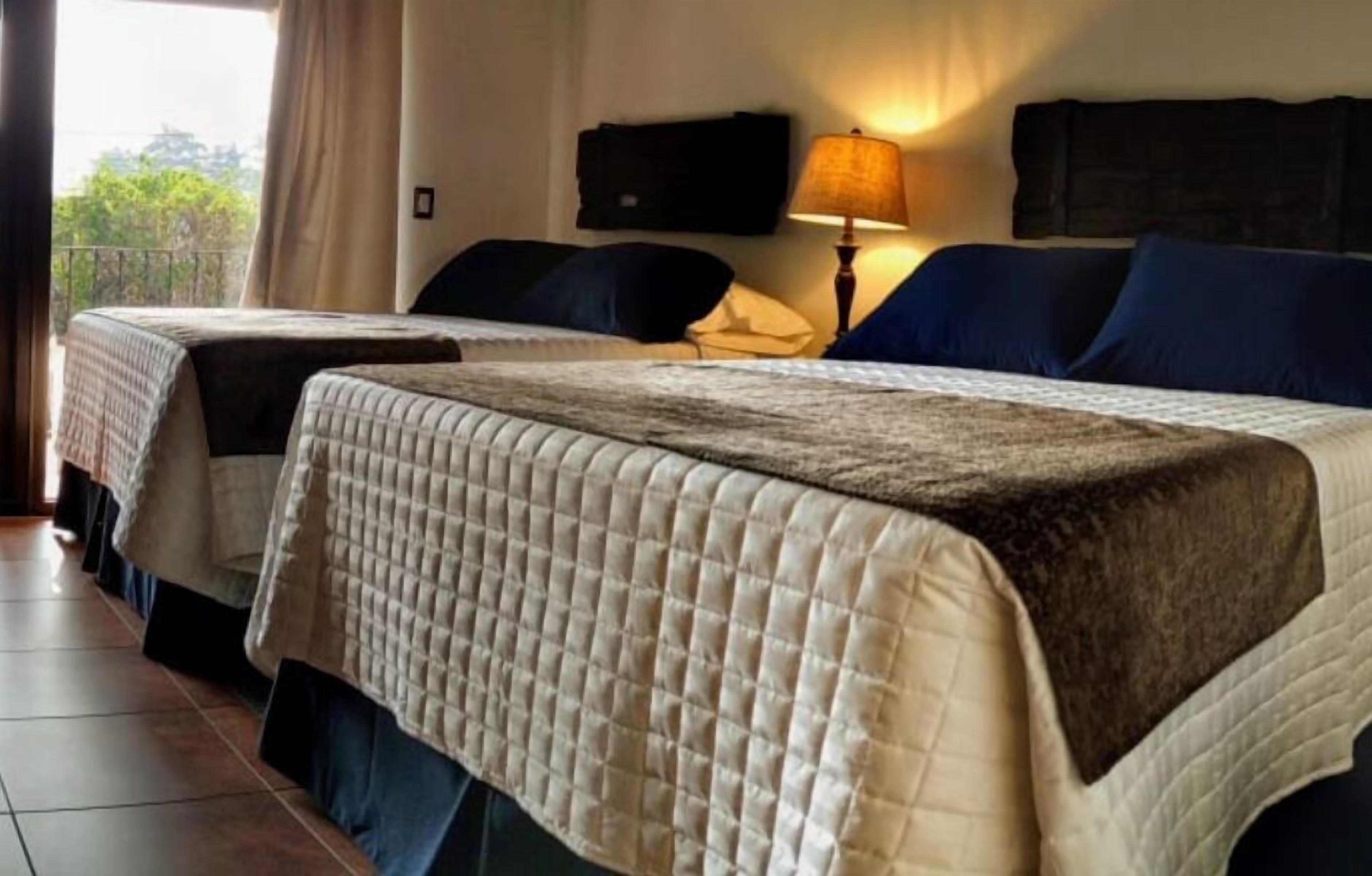 A hotel room with two beds, a lamp, and a view of greenery outside.