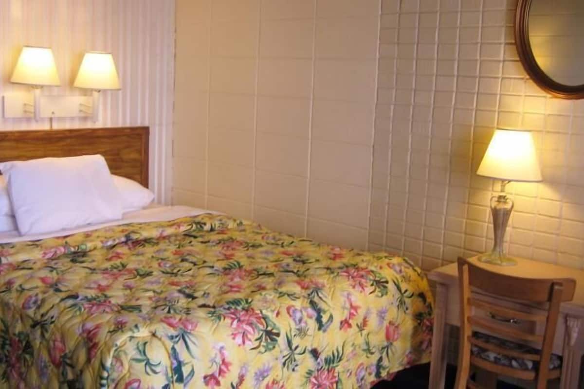 Una habitación de hotel con una cama, dos lámparas de noche, un cabecero de madera, una colcha con motivos florales, una silla de madera y un espejo montado en la pared.