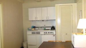Cocina funcional con armarios blancos, estufa y lavamanos. Hay una mesa con sillas y una lámpara sobre una mesita auxiliar.