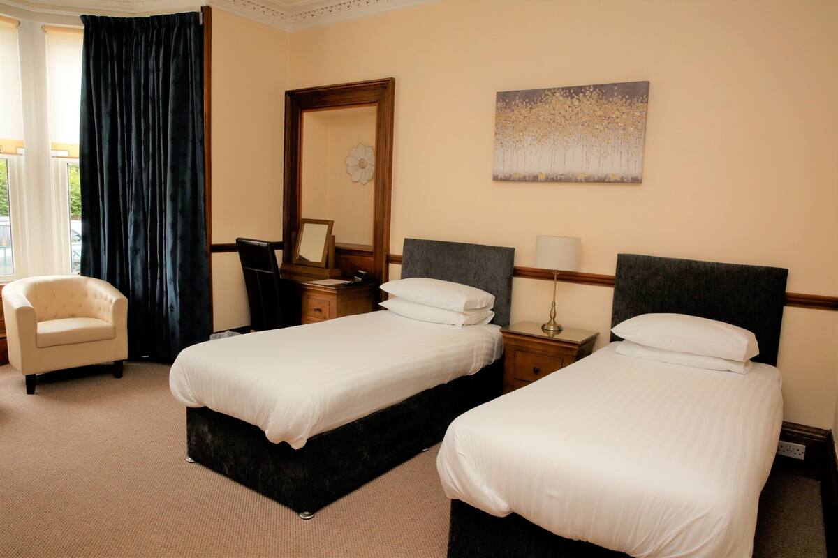A hotel room with two single beds, a chair, a desk, a mirror, and a painting on the wall.
