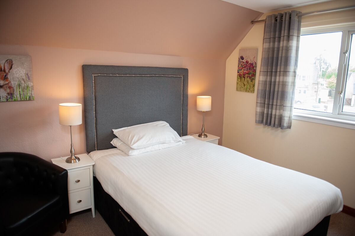A neatly made bed with a grey headboard, two bedside lamps, and a window with curtains.