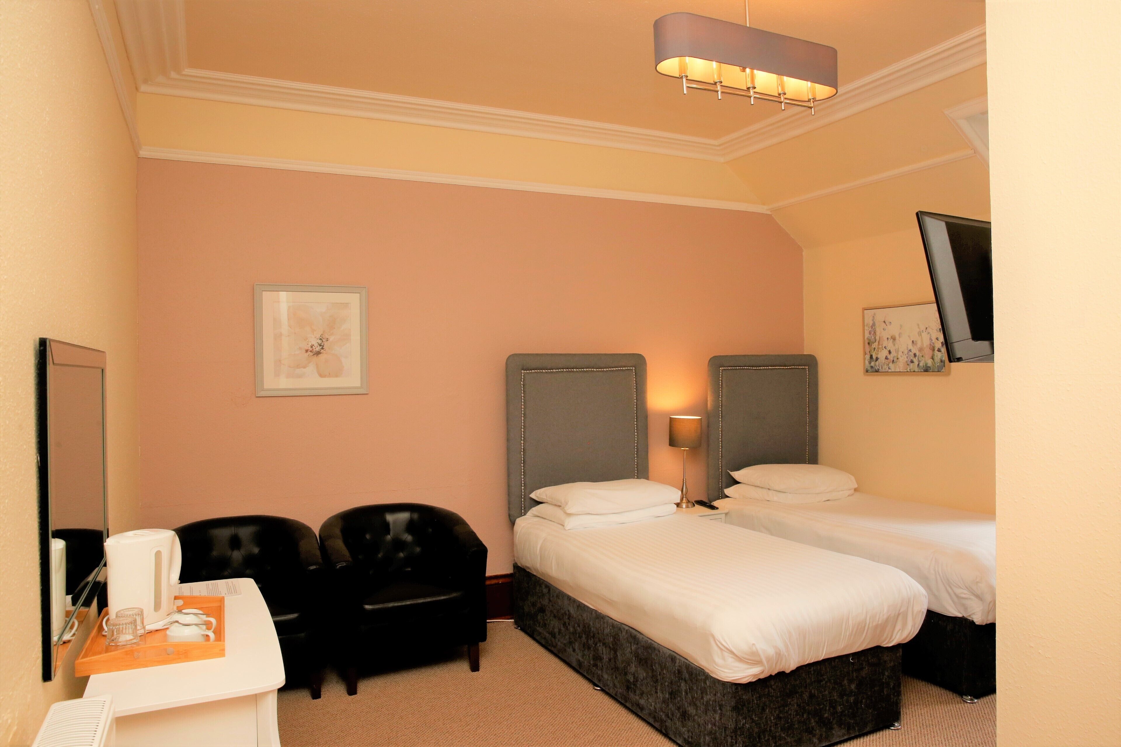 A hotel room with two beds, a desk with a kettle, a chair, and a flat-screen TV mounted on the wall.