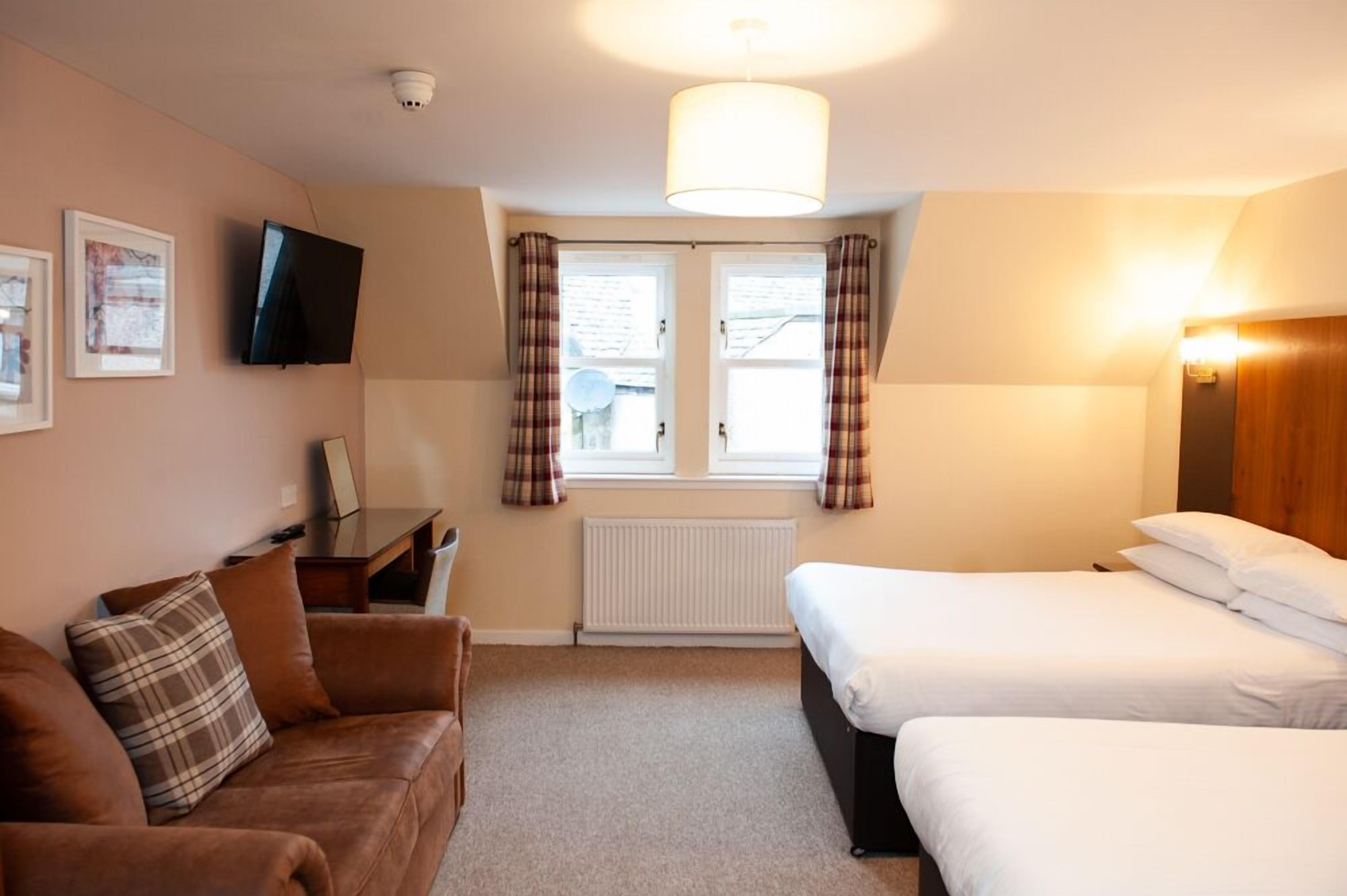 Een hotelkamer met een bruine leren bank, een bed met witte beddengoed, een aan de muur bevestigde televisie en een raam met geruite gordijnen.