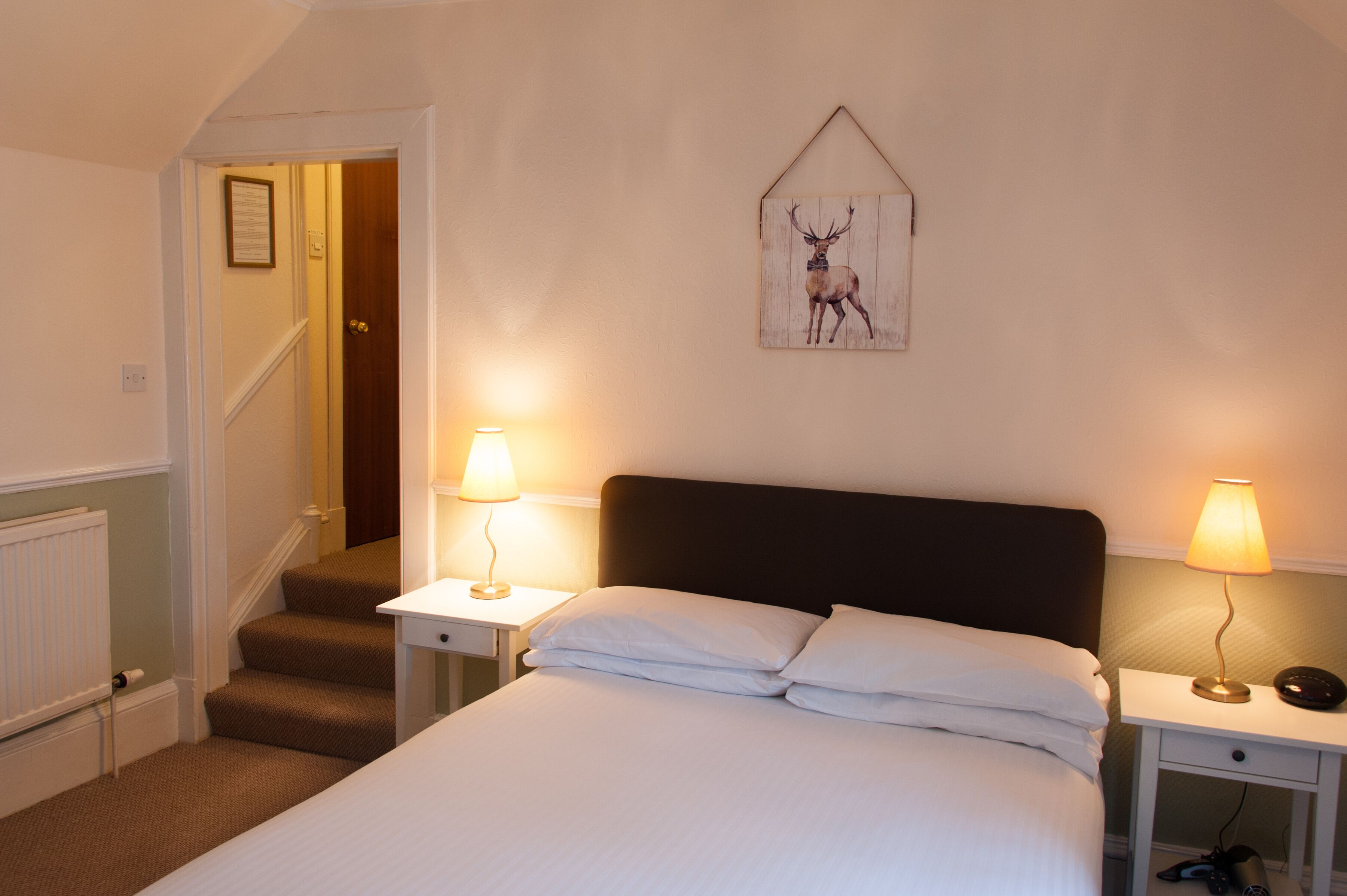 Une chambre à coucher avec un lit, deux tables de chevet avec des lampes, un tableau représentant un cerf encadré au mur et un escalier menant à une autre pièce.