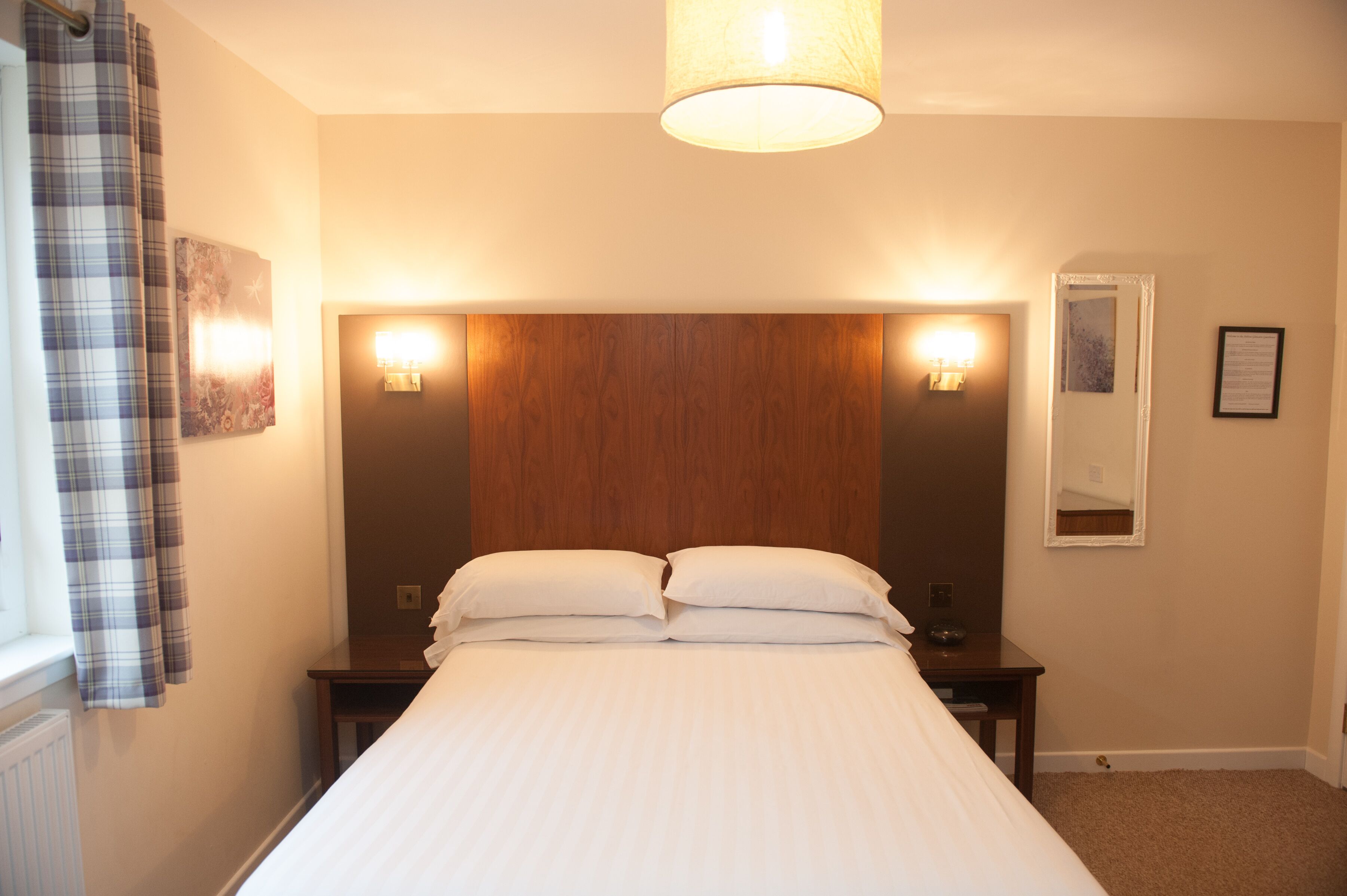 A neatly made bed with a wooden headboard, two bedside tables, a nightstand, a mirror, and a window with a plaid curtain.