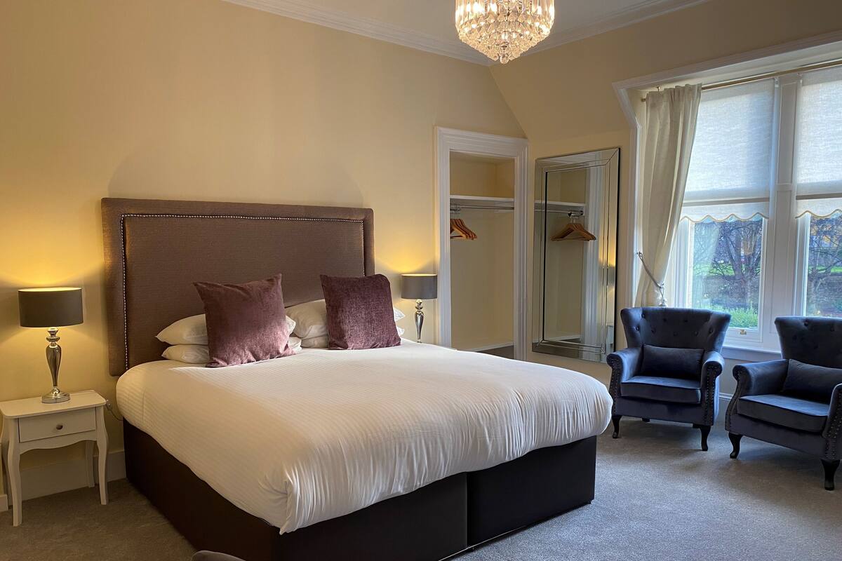A bedroom with a large bed, two bedside lamps, a mirror, a wardrobe, and two armchairs.