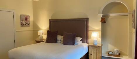 A neatly made bed with a headboard, two bedside tables with lamps, a small shelf with toiletries, and a door to the left.