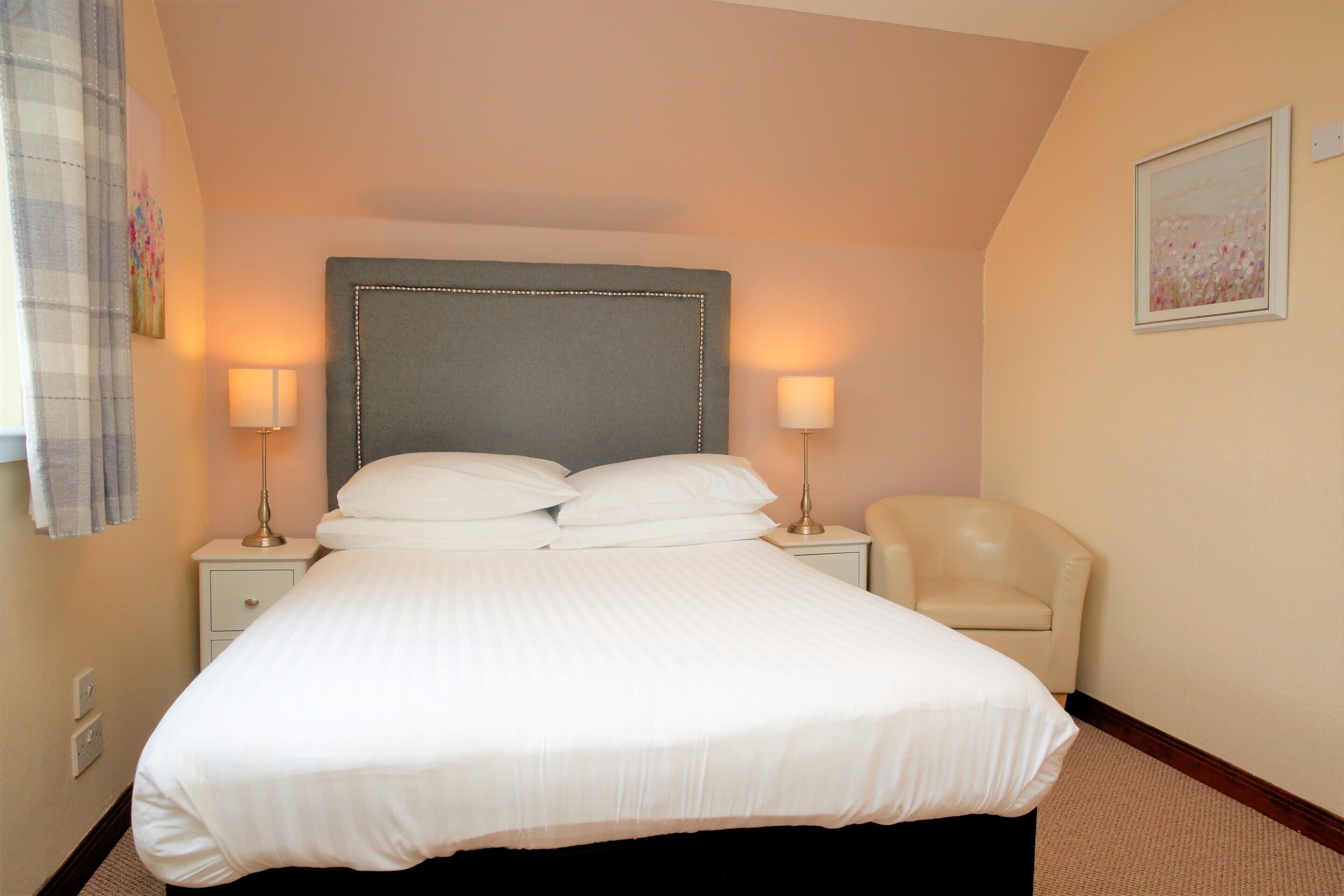 A neatly made bed with a grey headboard, two bedside lamps, a white chair, and a painting on the wall.