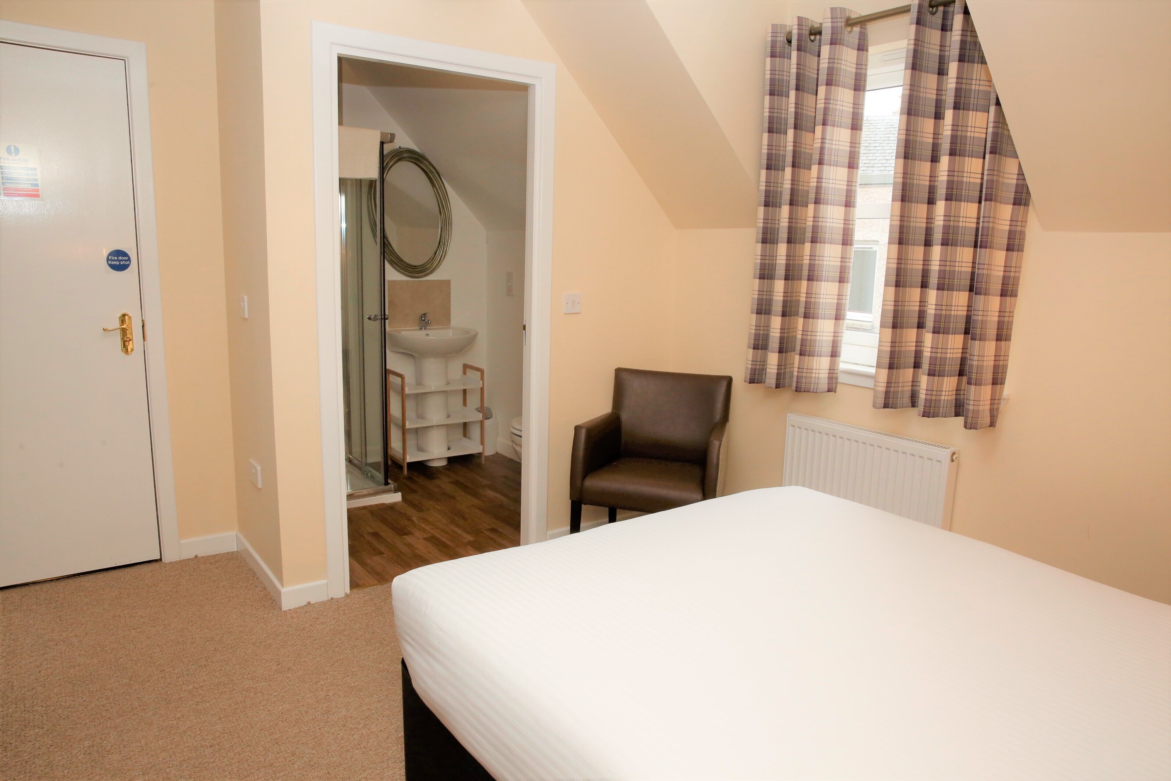 A bedroom with a bed, a chair, a bathroom with a sink and mirror, and a door.