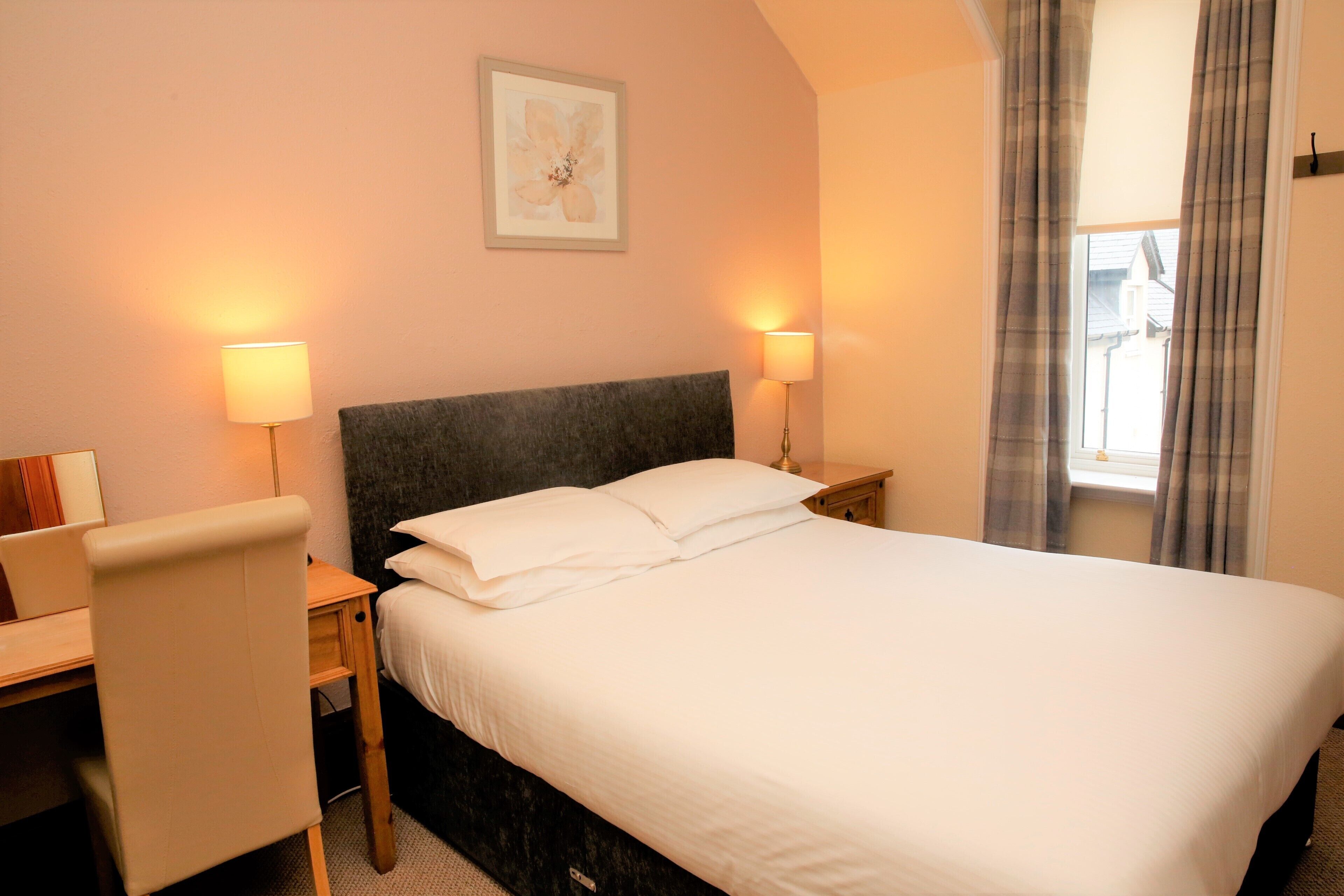 A hotel room with a bed, two bedside lamps, a desk, and a chair.