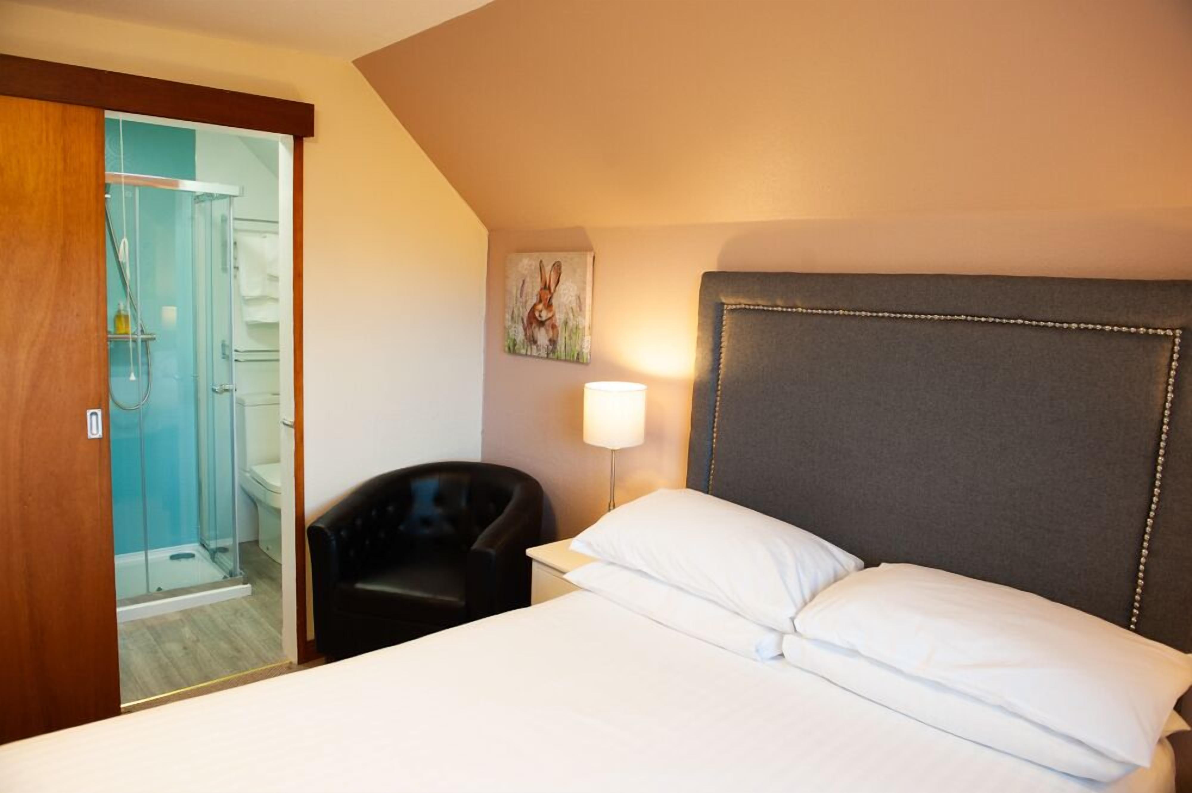 A hotel room with a bed, a chair, a bedside table with a lamp, a painting on the wall, and a shower area.