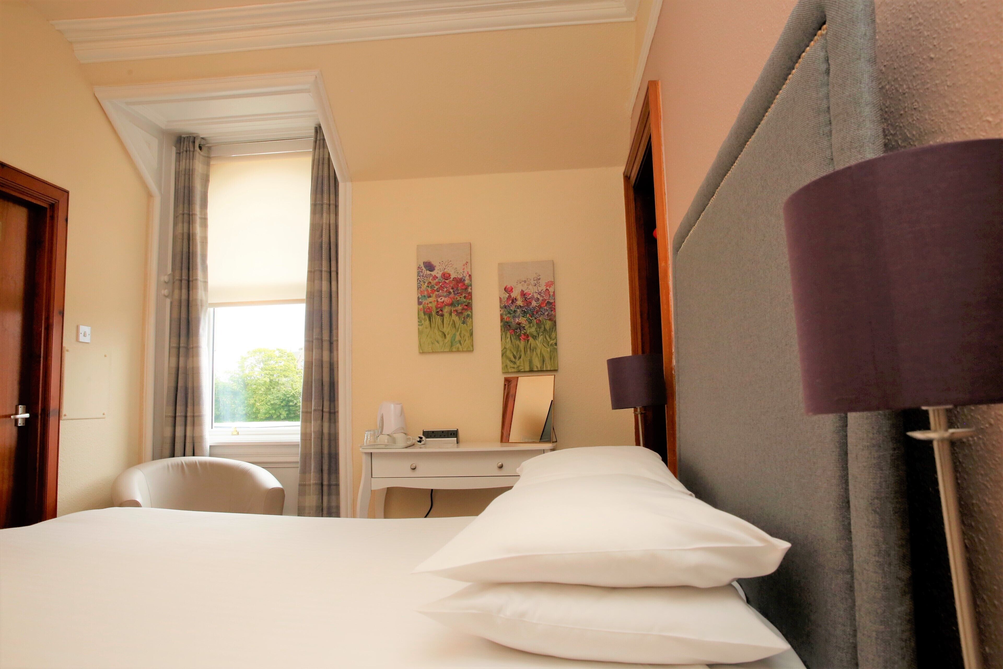 A hotel room with a bed, a desk, a chair, and two paintings on the wall.