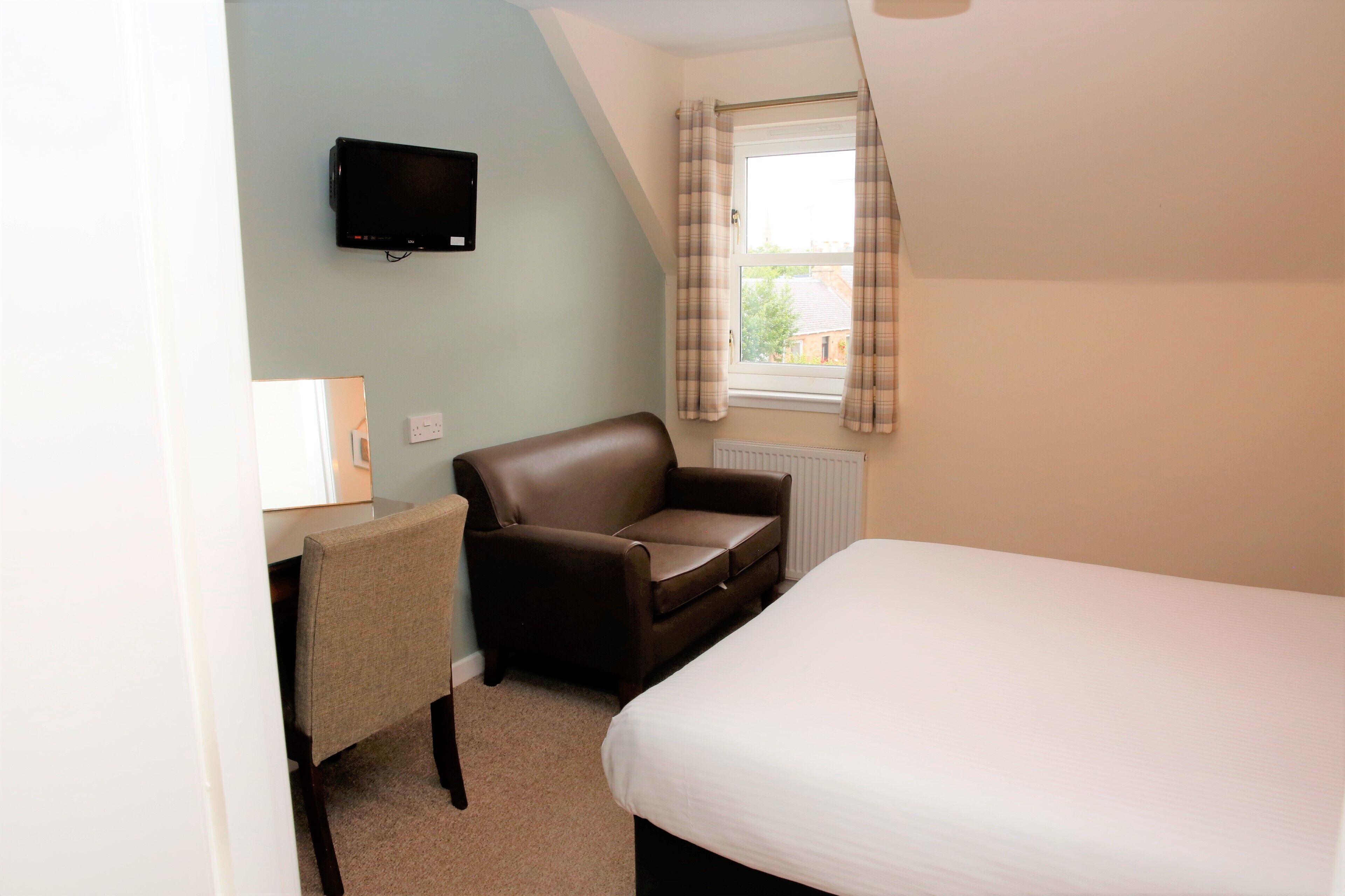 A small, well-lit room with a single bed, a brown leather chair, a small desk with a lamp, and a television mounted on the wall.