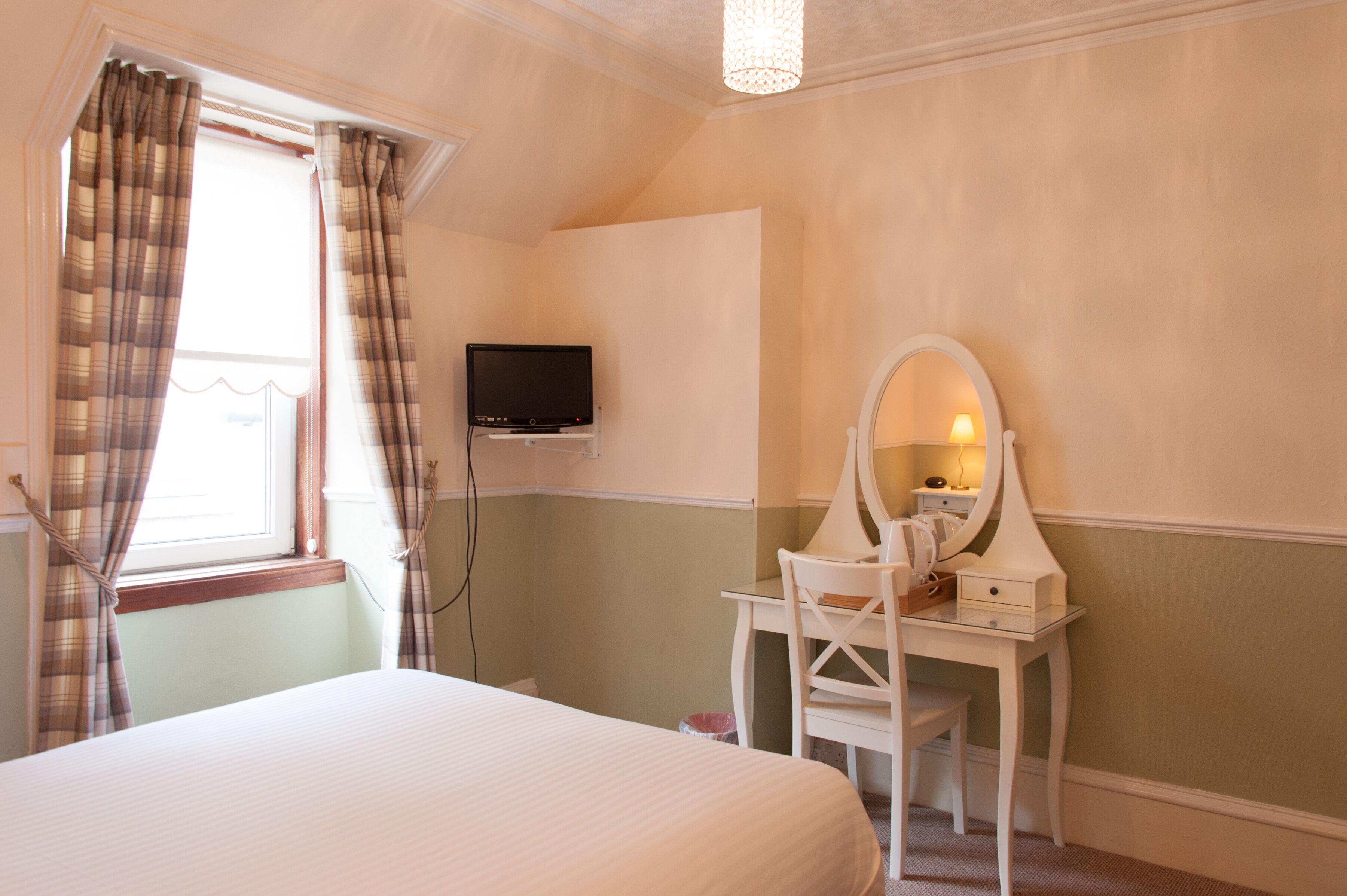 A hotel room with a bed, a desk with a mirror, a television on the wall, and a window with curtains.