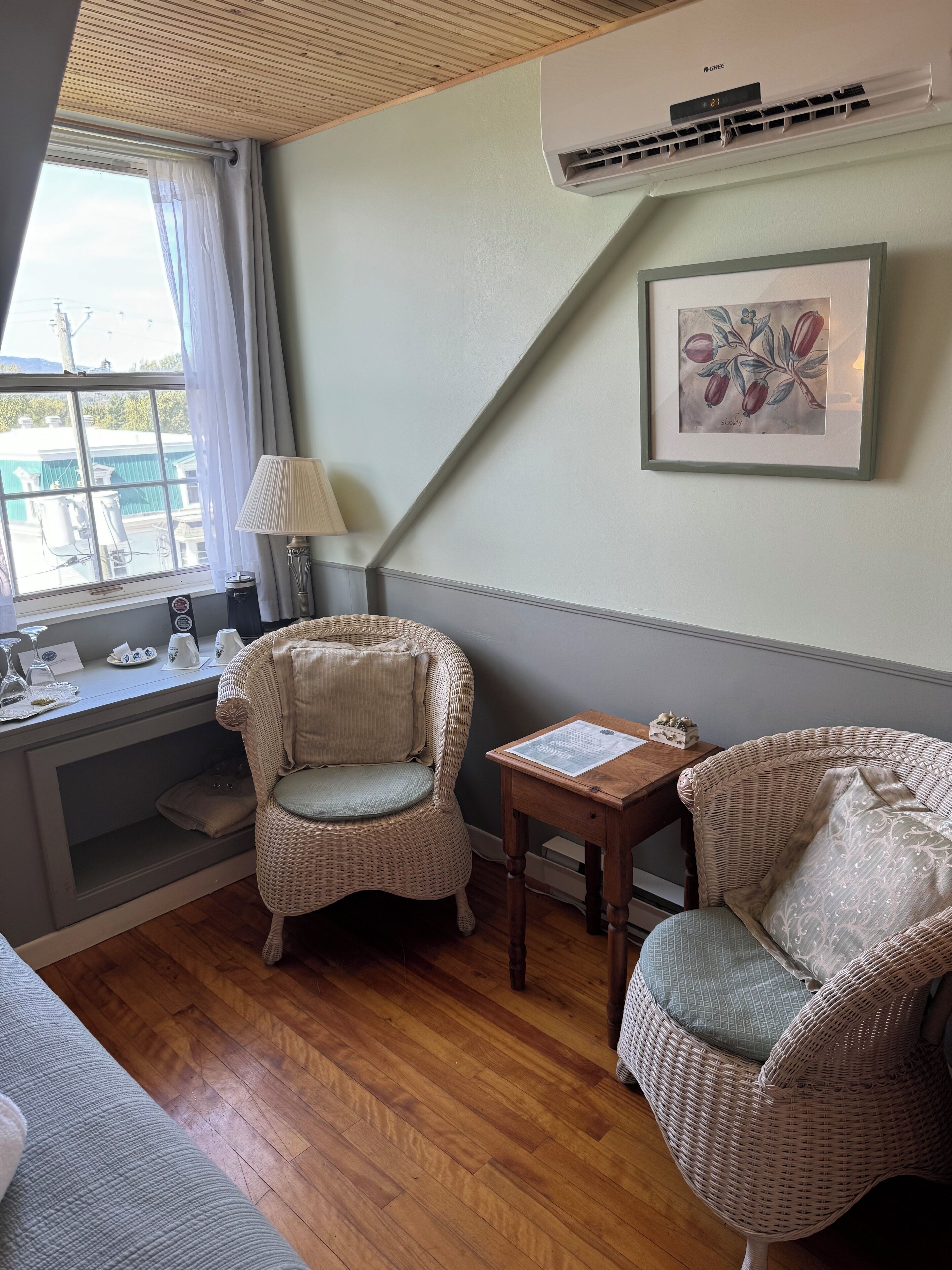 Una habitación acogedora con techo de madera, una ventana con cortinas, un sillón de mimbre, una mesita con una lámpara y un cuadro en la pared.